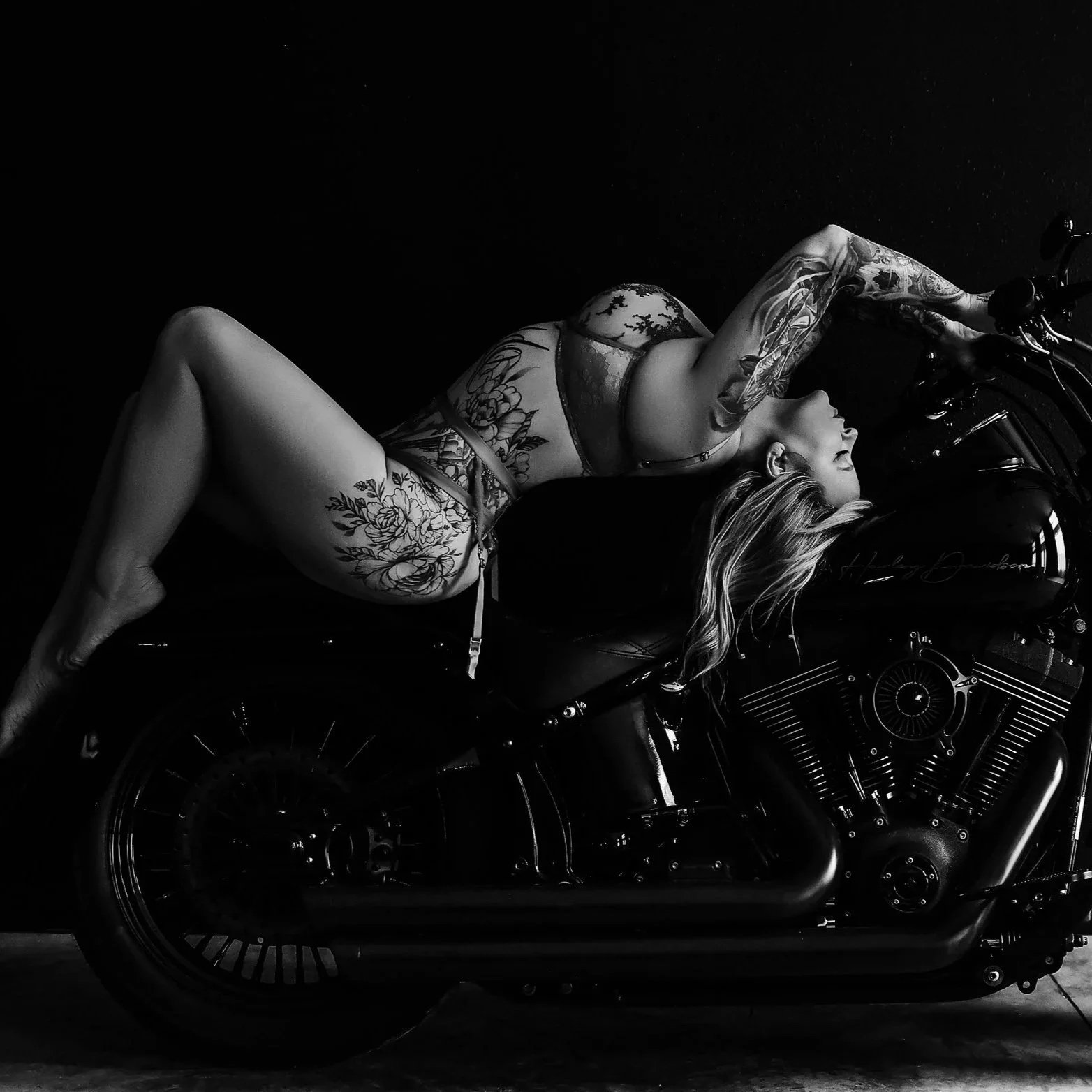 A woman with tattoos lying on a motorcycle, wearing lingerie in a black and white photograph.
