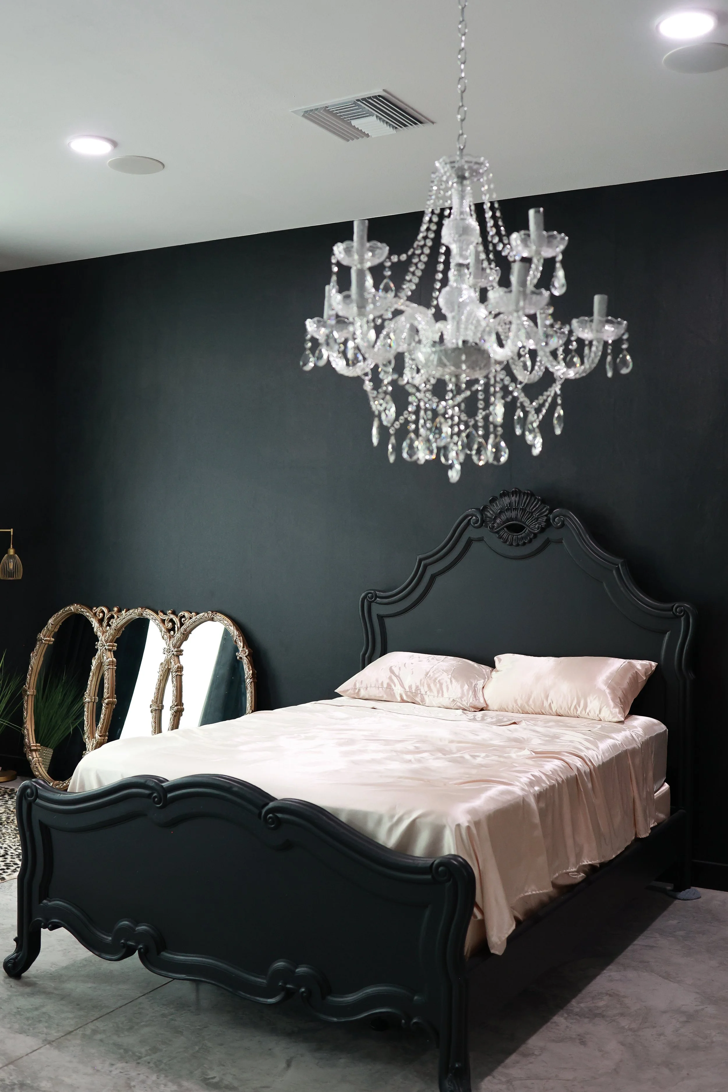 A bedroom with a black headboard, four-bead White satin sheets with matching pillows, a tall gold-framed mirror, and a crystal chandelier hanging from the ceiling. The room has a black wall and a grey floor.