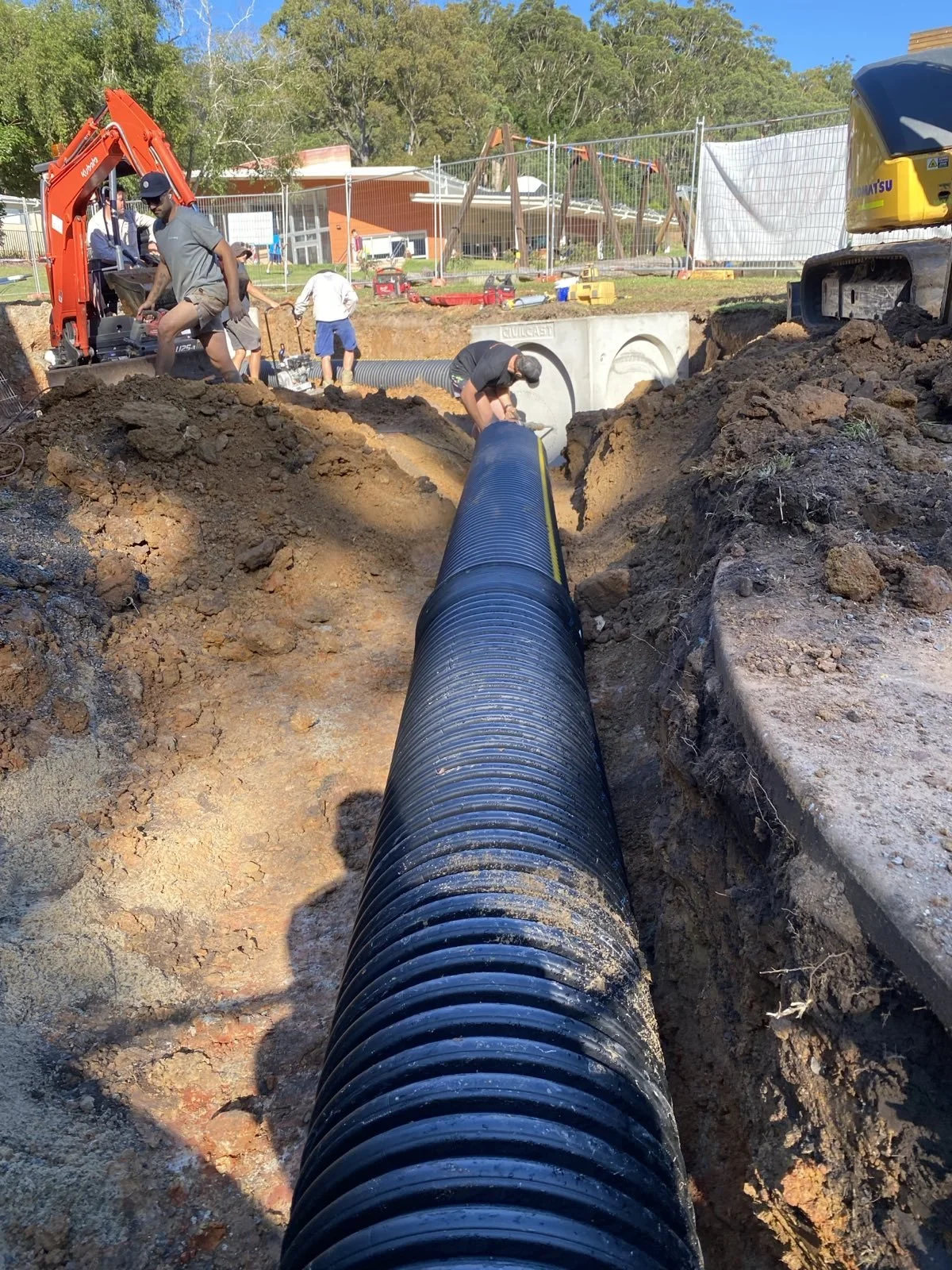 Construction workers laying down a large black pipe in a trench at an outdoor construction site with equipment and machinery around.