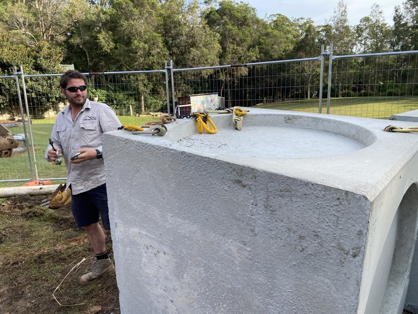 A man wearing sunglasses, a light-colored shirt, and dark shorts stands next to a large concrete structure outdoors, with tools placed on top of the structure, against a background of trees and a grassy field.