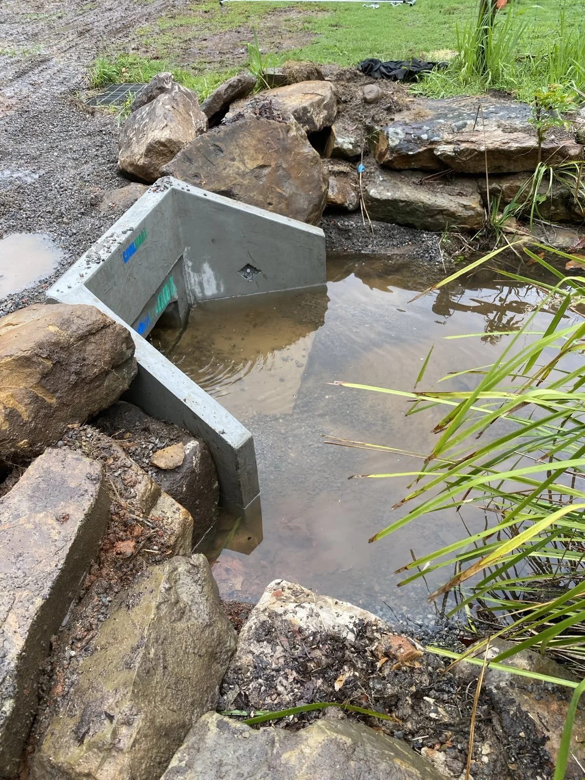 A yard with a pond area, large rocks surrounding it, and a rectangular concrete structure partially submerged in the water.