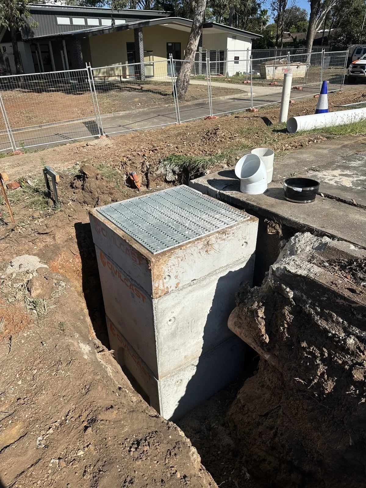 Underground utility access box next to construction site with pipes and drainage components near a sidewalk and residential houses.