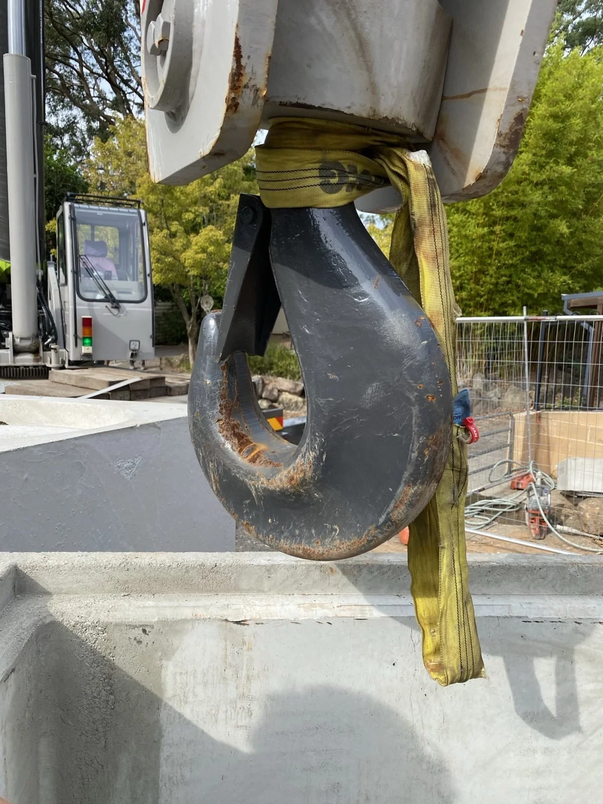Close-up of a large hook hanging from a crane hook, with a yellow strap attached, and a construction site in the background.