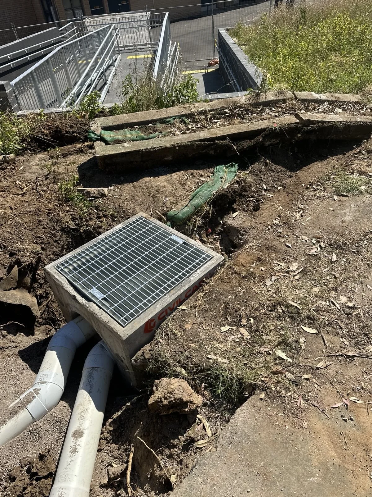 Underground utility box with two white pipes connected to it, surrounded by dirt and gravel, with some green plants and construction debris nearby. A set of metal railings and a staircase are visible in the background.