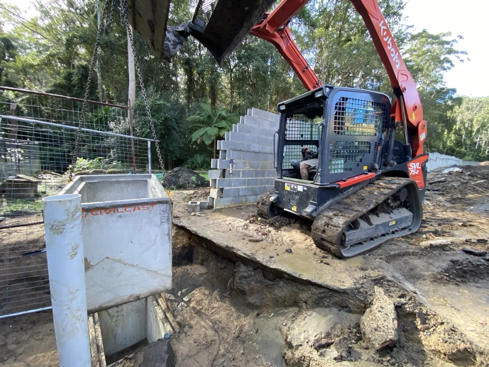 Construction site with a mini excavator moving dirt, a cinder block wall, and a fenced area with trees in the background.