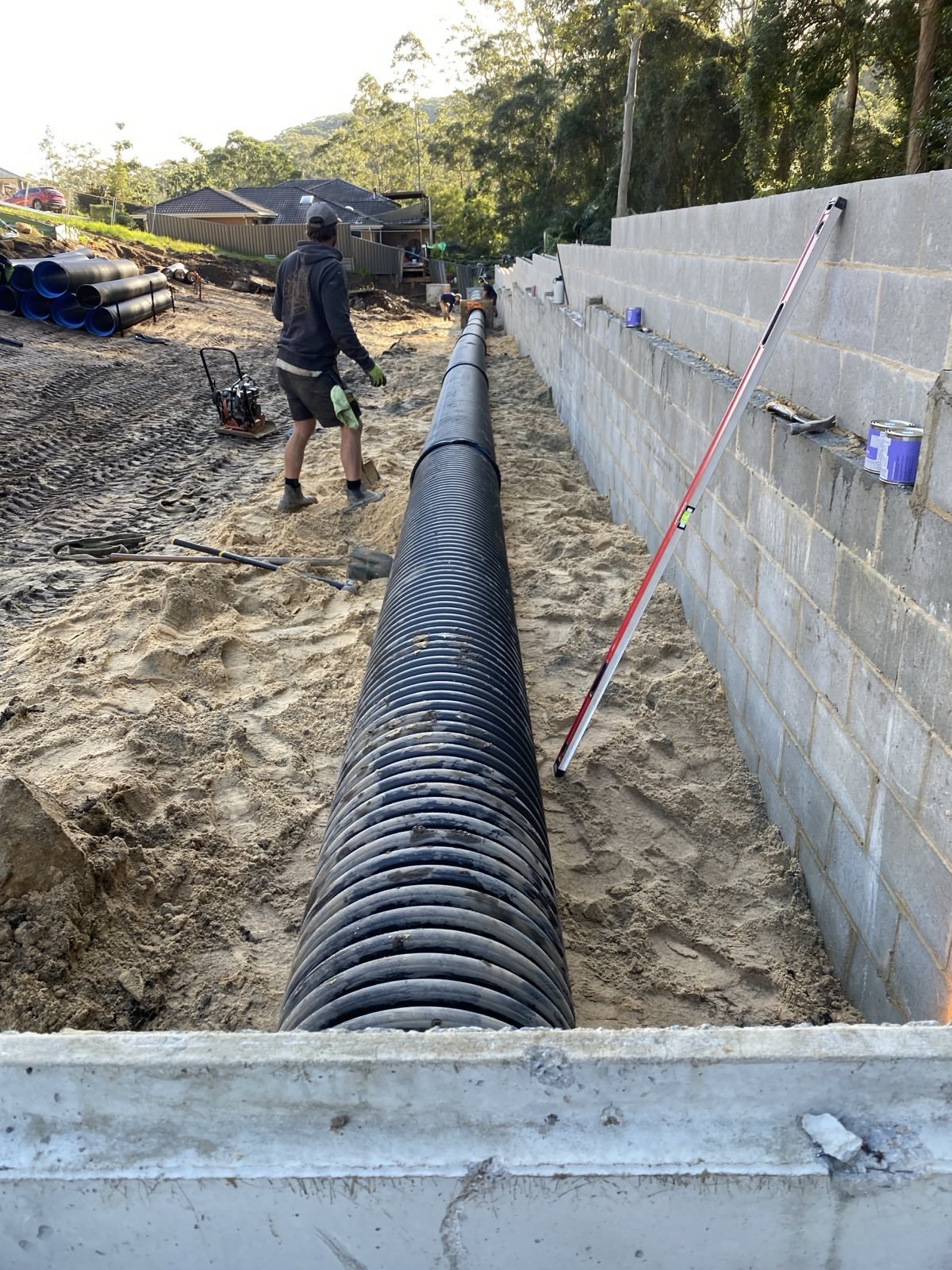 Construction worker installing a large black corrugated pipe along a dug trench adjacent to a concrete block wall with cans of paint and tools nearby.