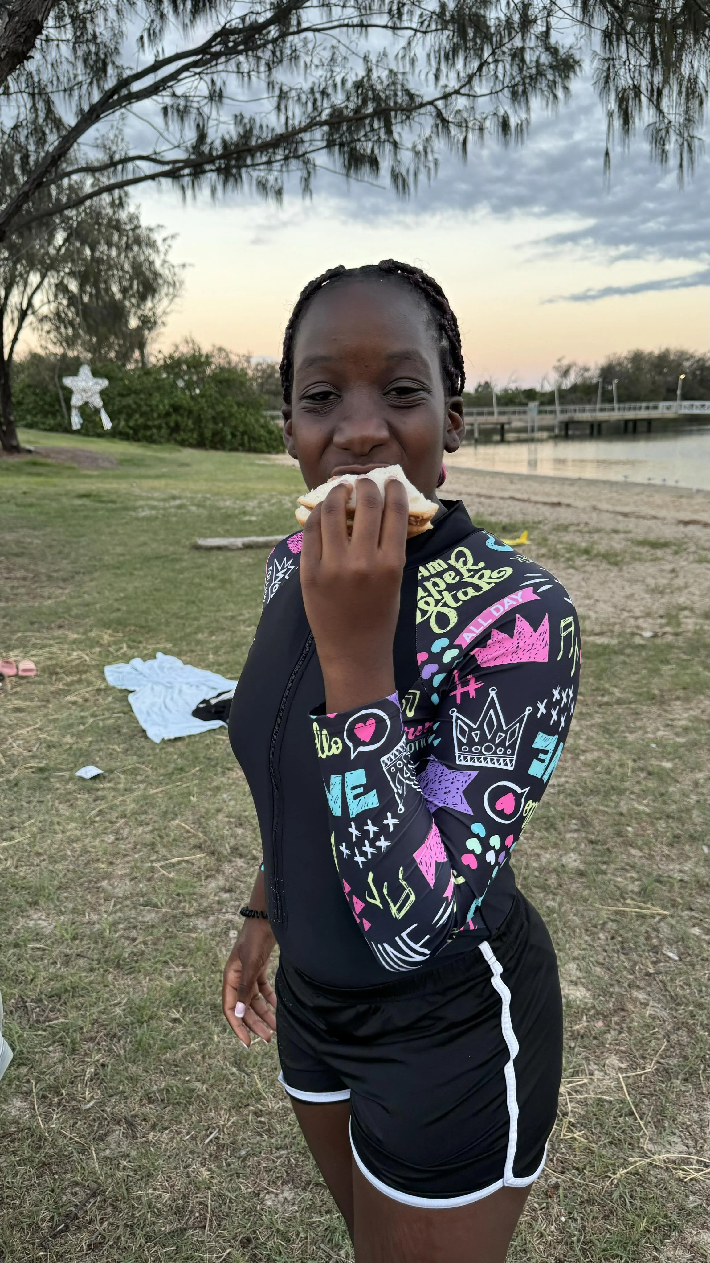 A young girl with braided hair is outdoors, holding a sandwich and appearing to eat it, with a lake and trees in the background during sunset.