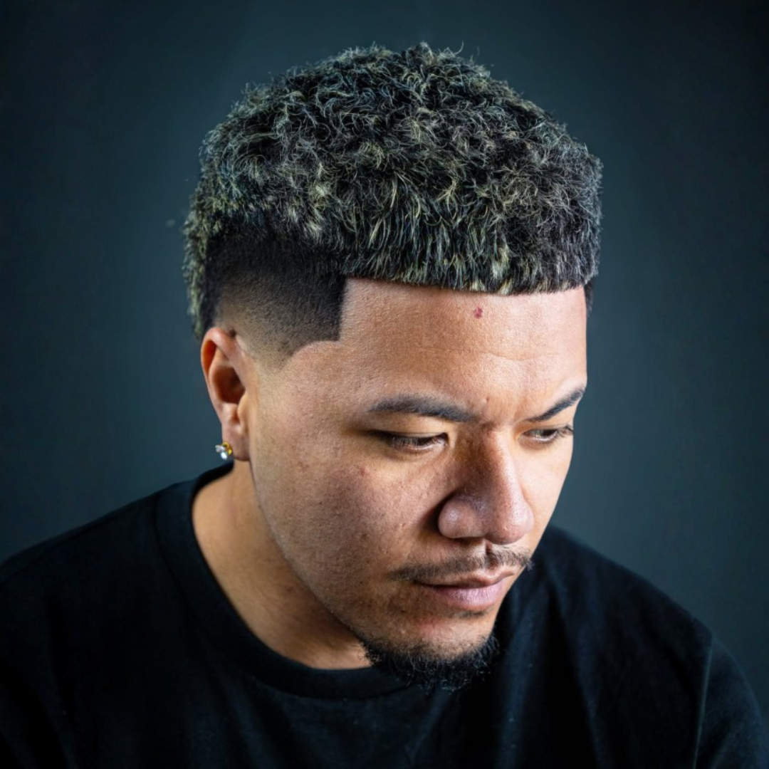 Close-up of a young man with short, curly dark hair with blonde highlights, wearing a black shirt, looking downward against a dark background.
