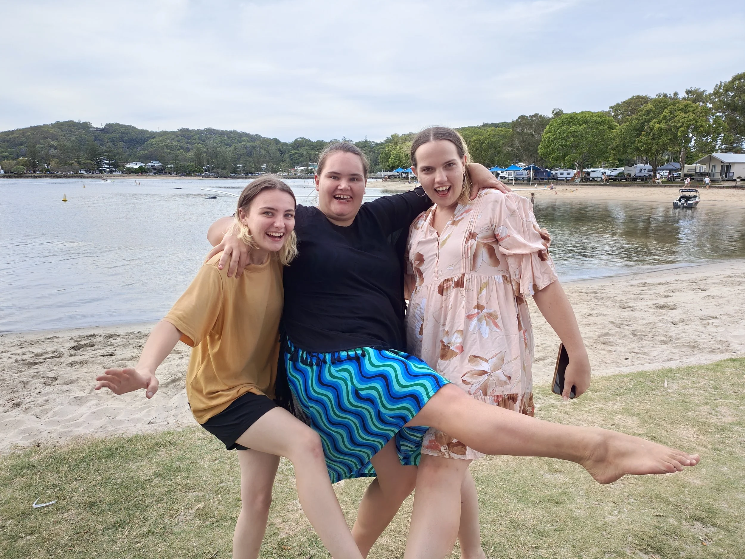 Three women smiling and posing together on a beach, with water, trees, and houses in the background.