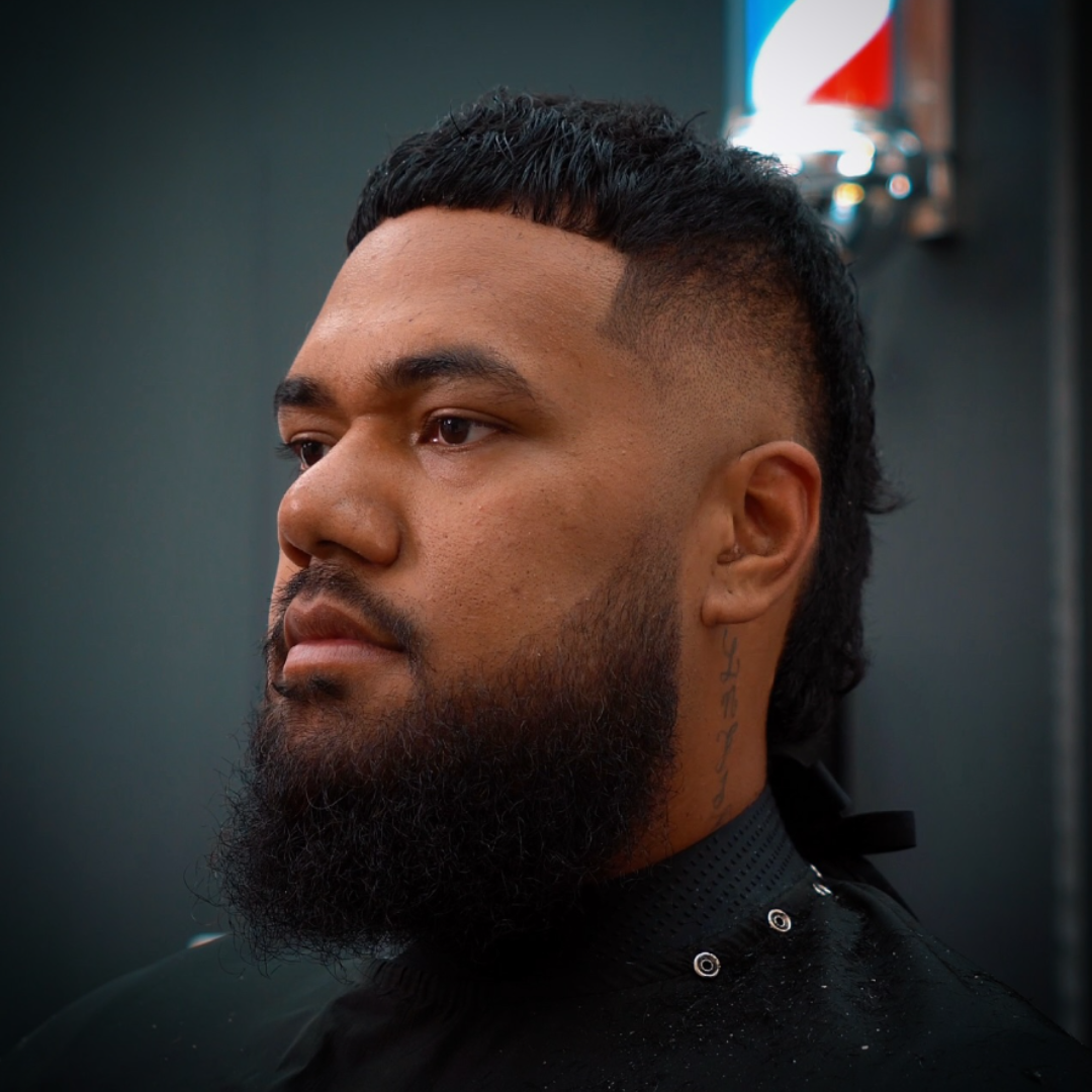 A man with a beard and a fresh haircut, featuring a fade on the sides and short curly hair on top, poses in a barber shop with a Pepsi sign in the background.