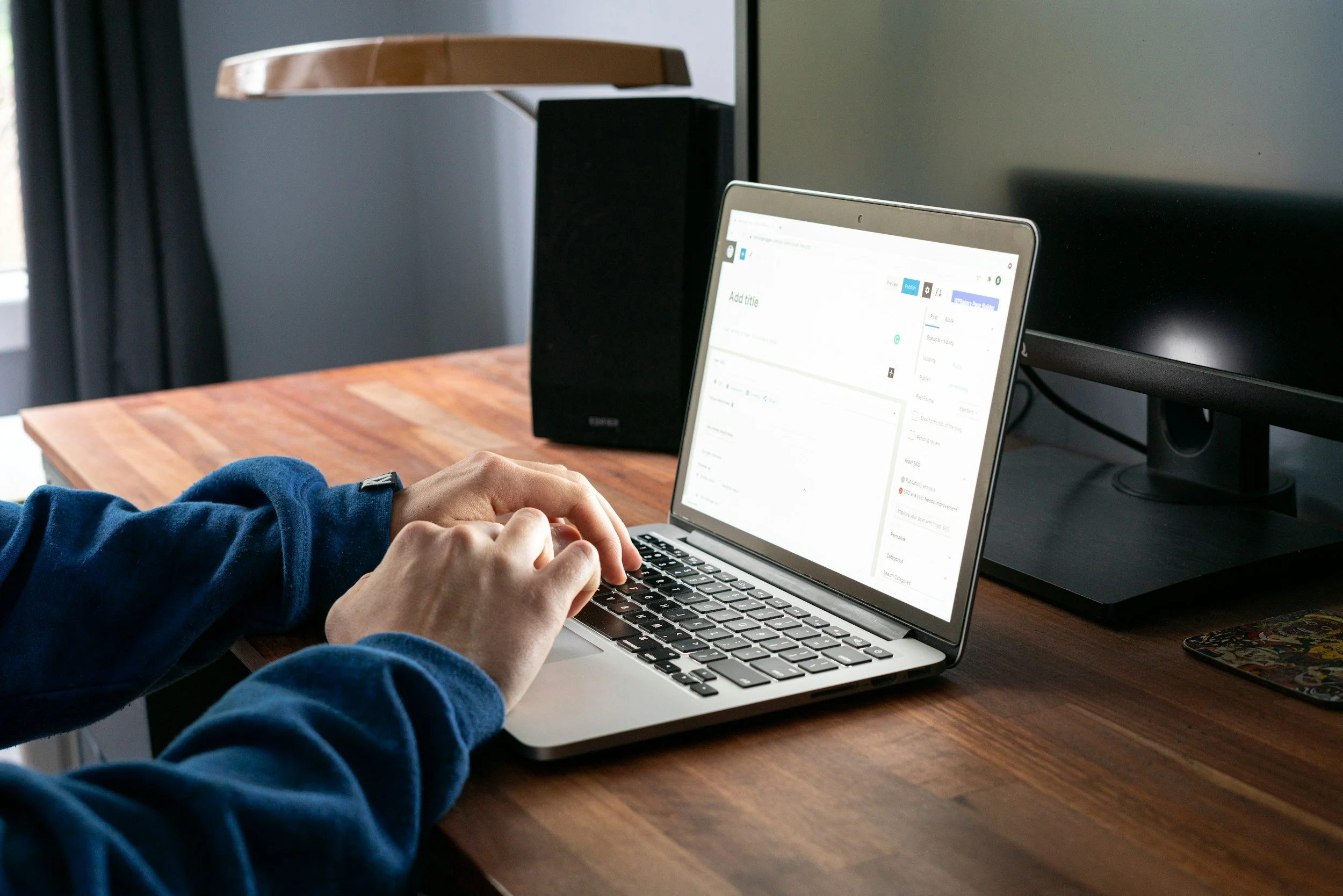 Person using a laptop at a wooden desk, with a monitor and speaker in the background.