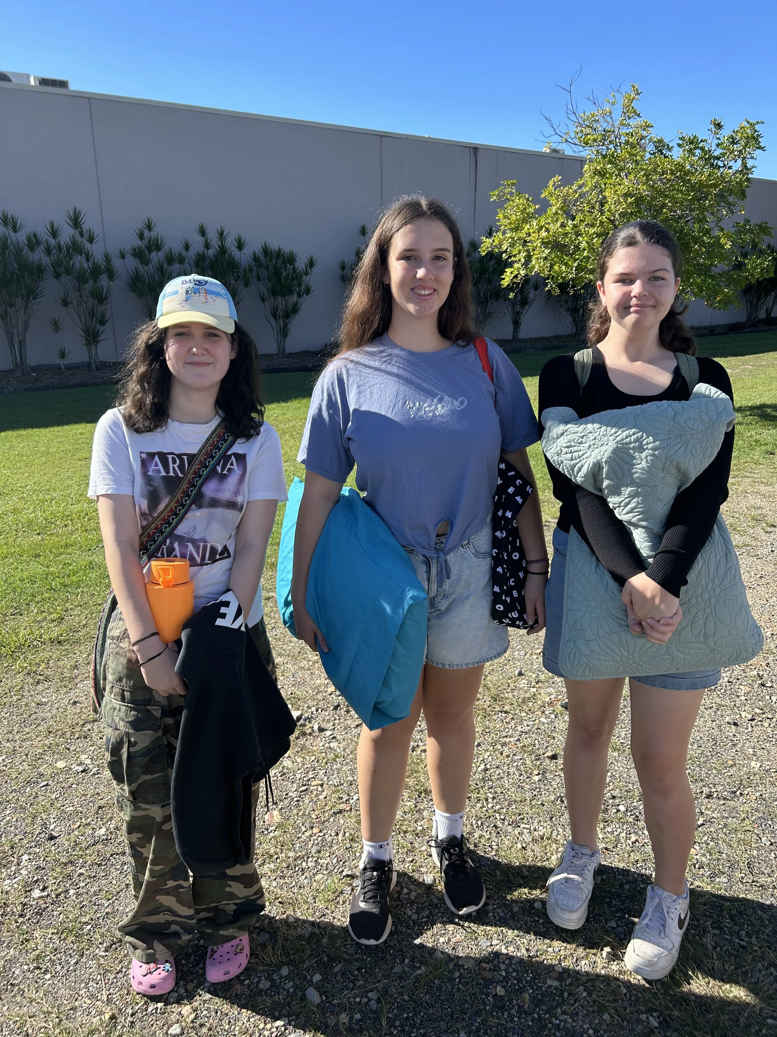 Three young girls standing outdoors on a sunny day, each holding or carrying bags or water bottles, with a green lawn, trees, and a light gray wall in the background.