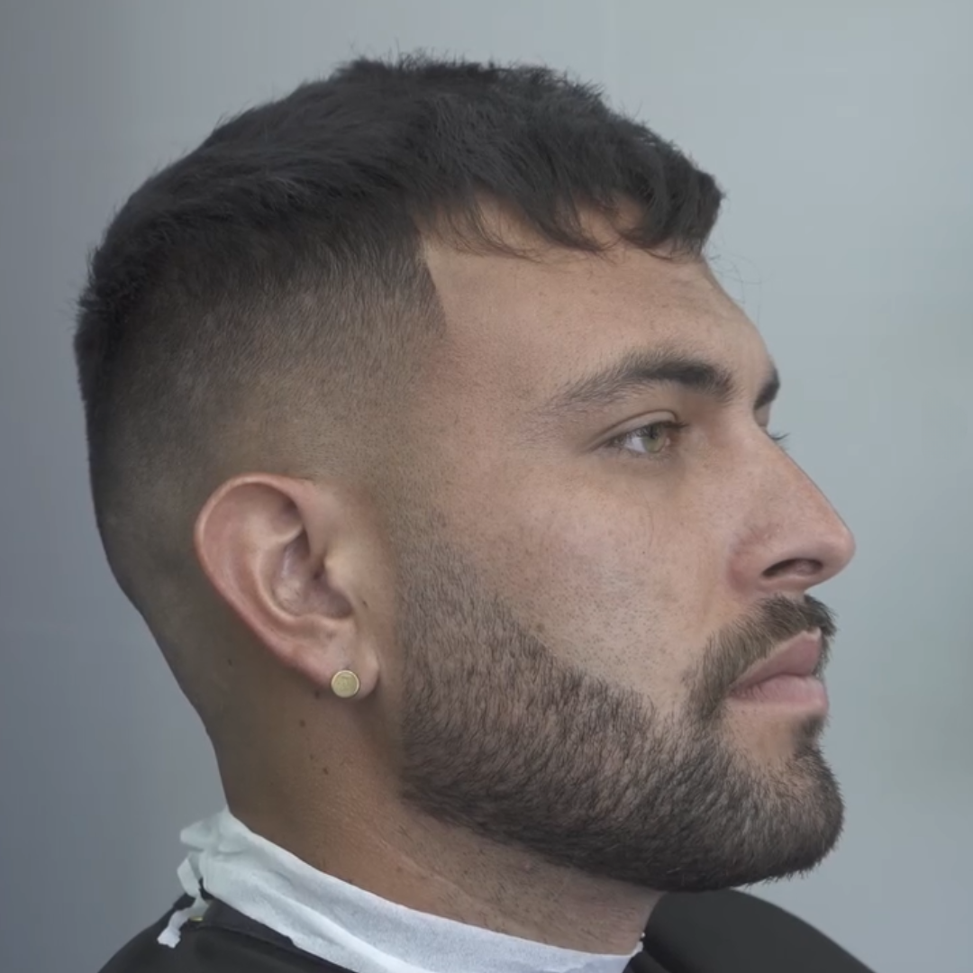 Side profile of a man with a freshly cut hairstyle, beard, and wearing a small earring.