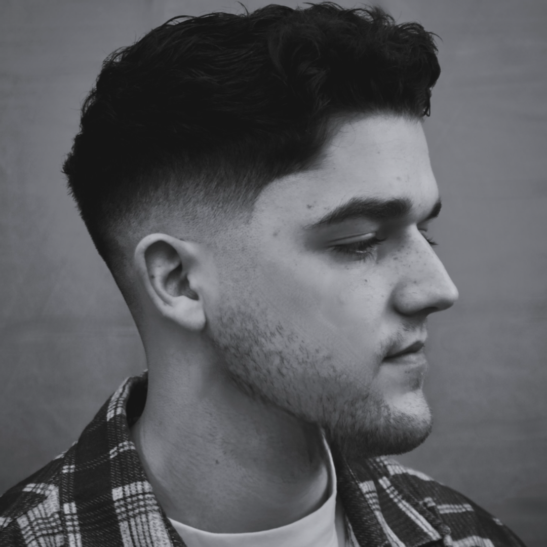 Black and white close-up of a young man with short, wavy hair, facial hair, and a plaid shirt, in profile view against a plain background.