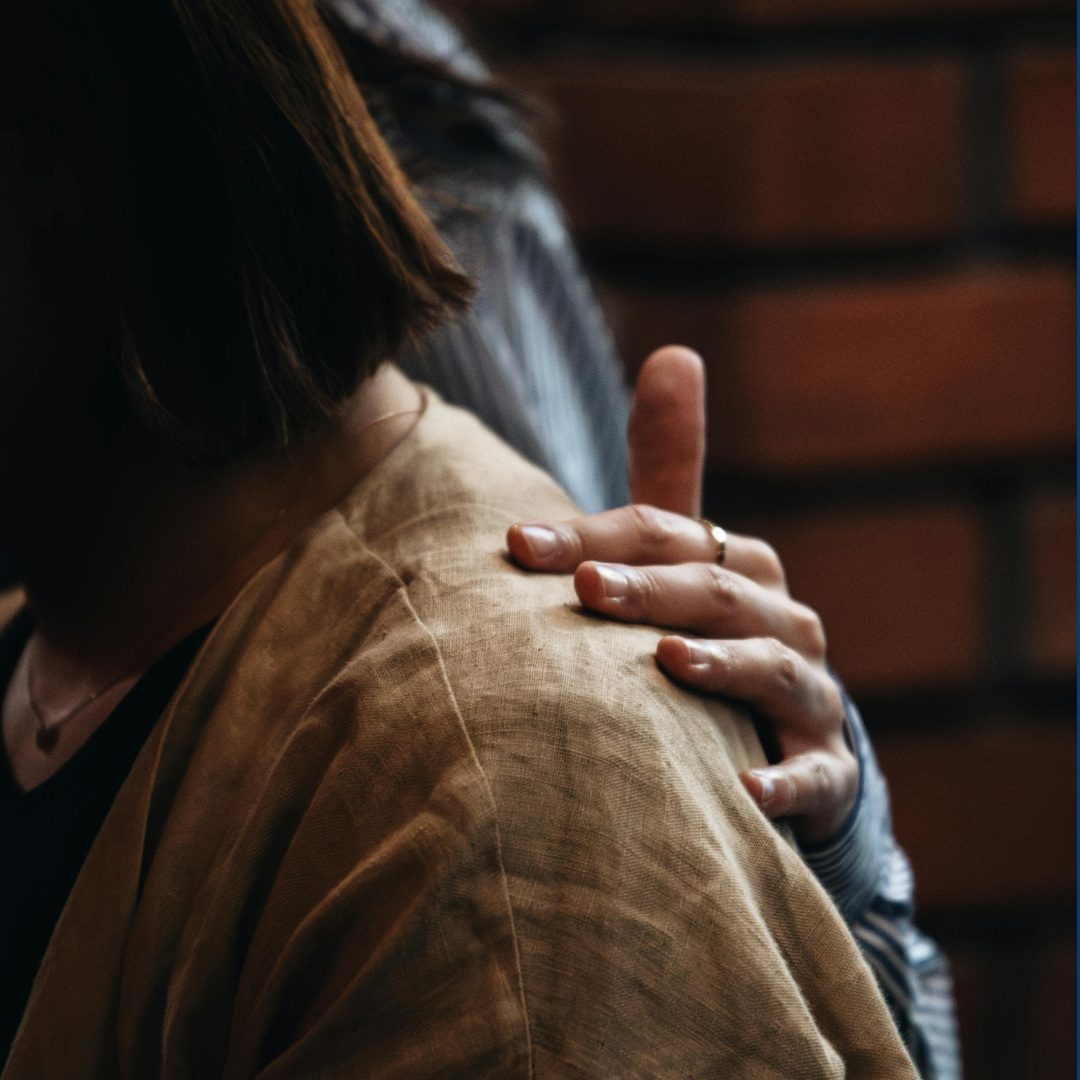 Close-up of a person with shoulder-length brown hair touching their shoulder with their hand, wearing a beige jacket and a ring, with a background of brick wall.