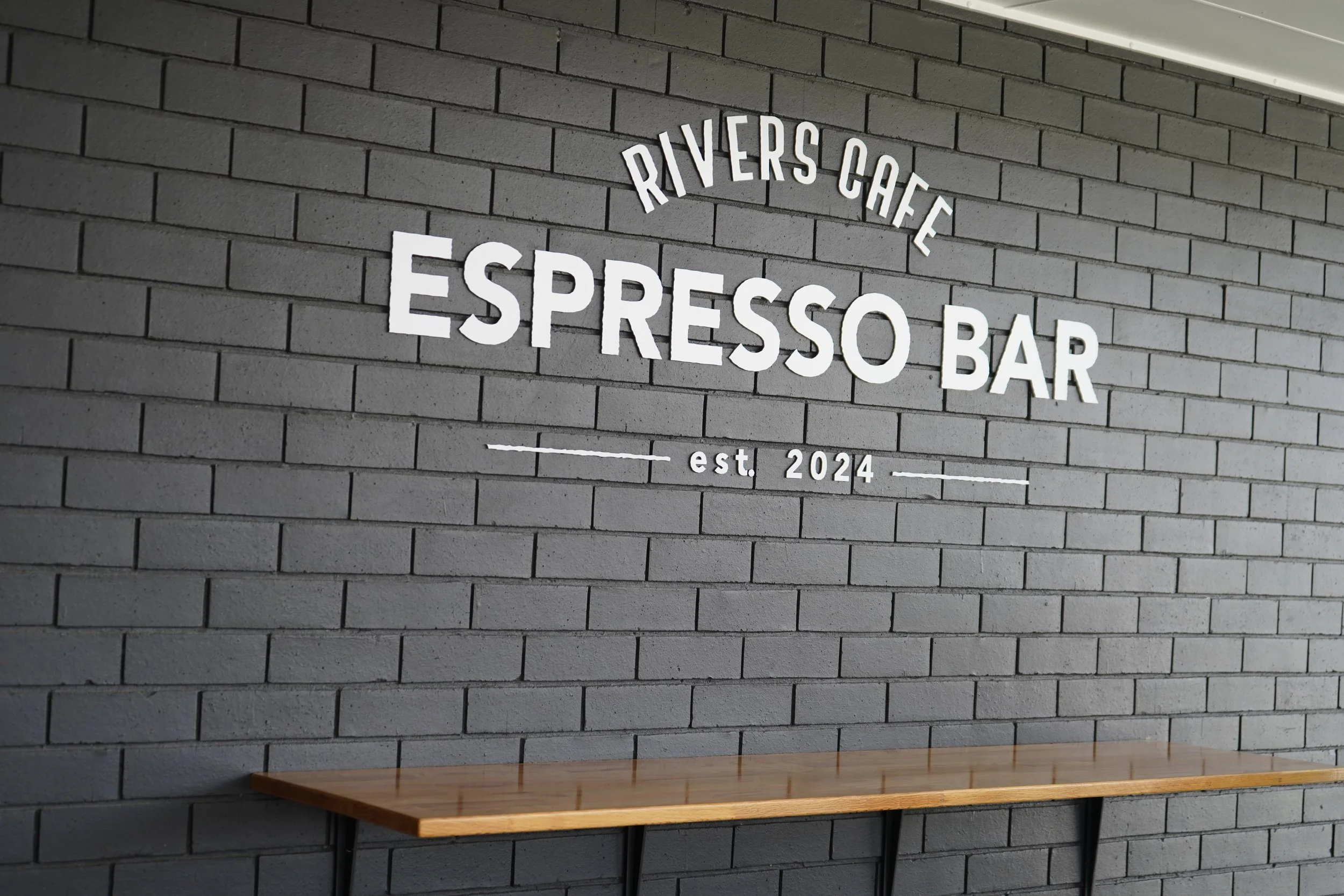Interior of a cafe with a gray brick wall featuring a sign that reads 'Rivers Cafe Espresso Bar Est. 2024' in white letters. There is a wooden table below the sign.
