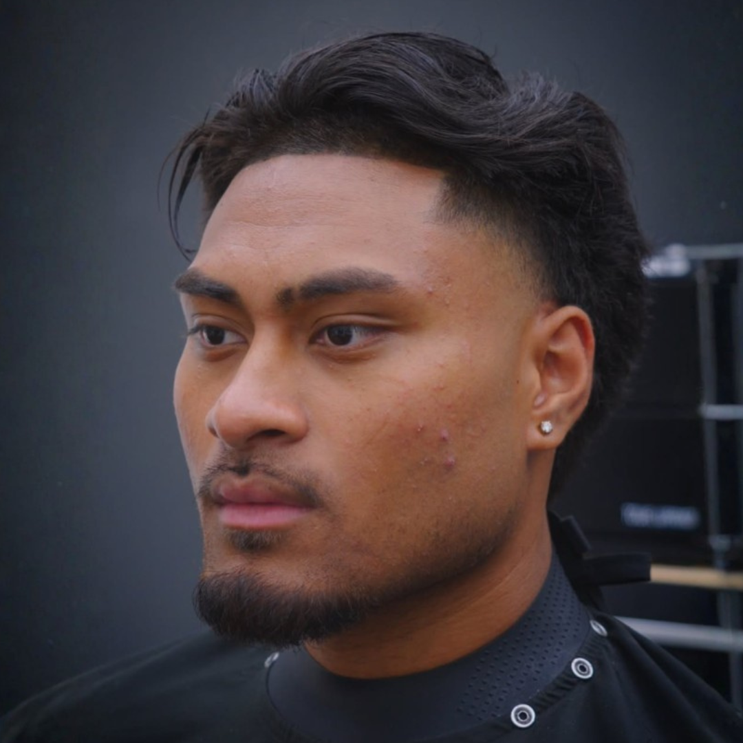A close-up of a young man with dark hair and a goatee, wearing a black shirt with a textured collar and small earring, against a dark background.