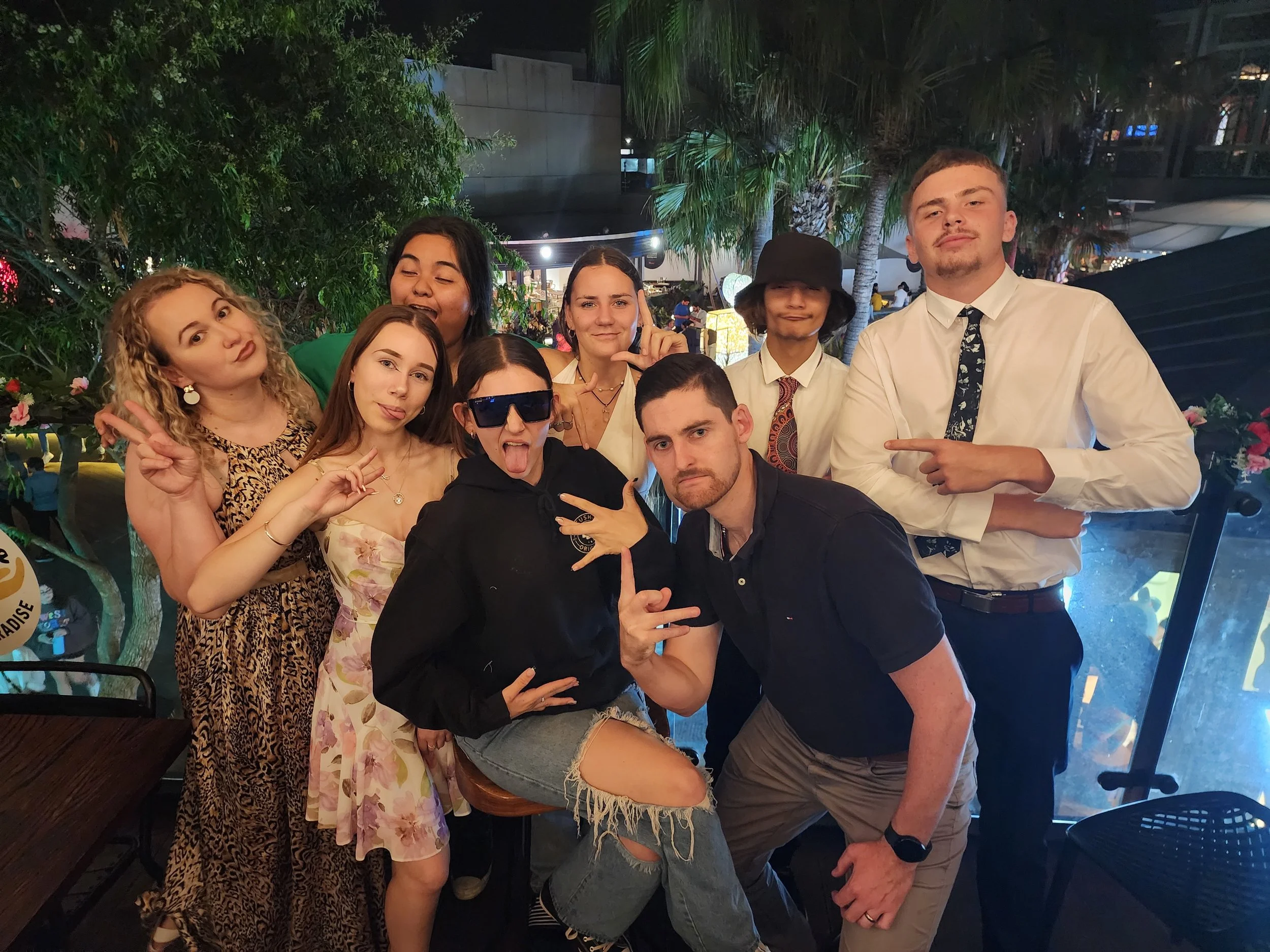 A group of nine young adults posing together outdoors at night. They are smiling and making various gestures, with some wearing trendy clothing and accessories. The background features palm trees and decorative lighting.