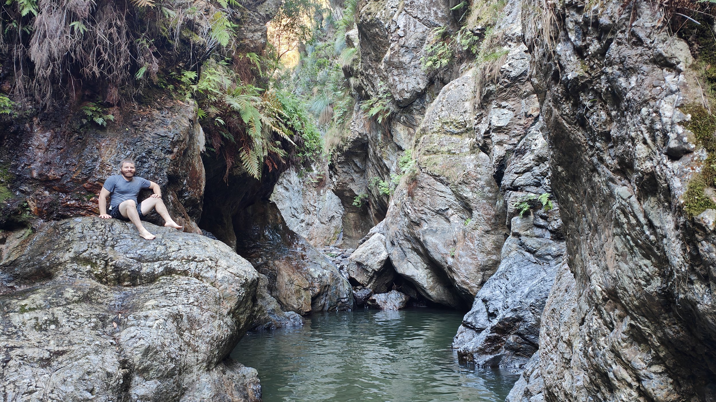 A man sitting on a large rock in a narrow canyon with water at the bottom, surrounded by steep rocky walls and lush foliage.