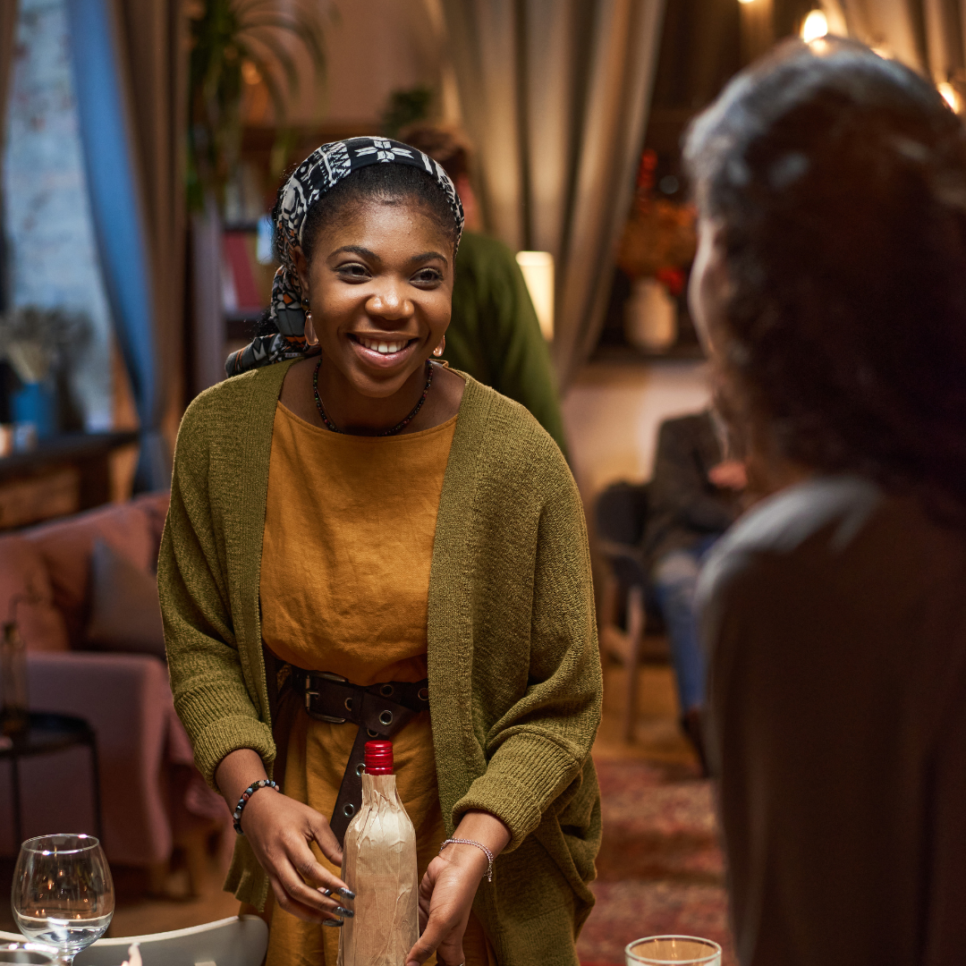 A woman smiling and holding a bottle at a social gathering in a warmly lit indoor setting.