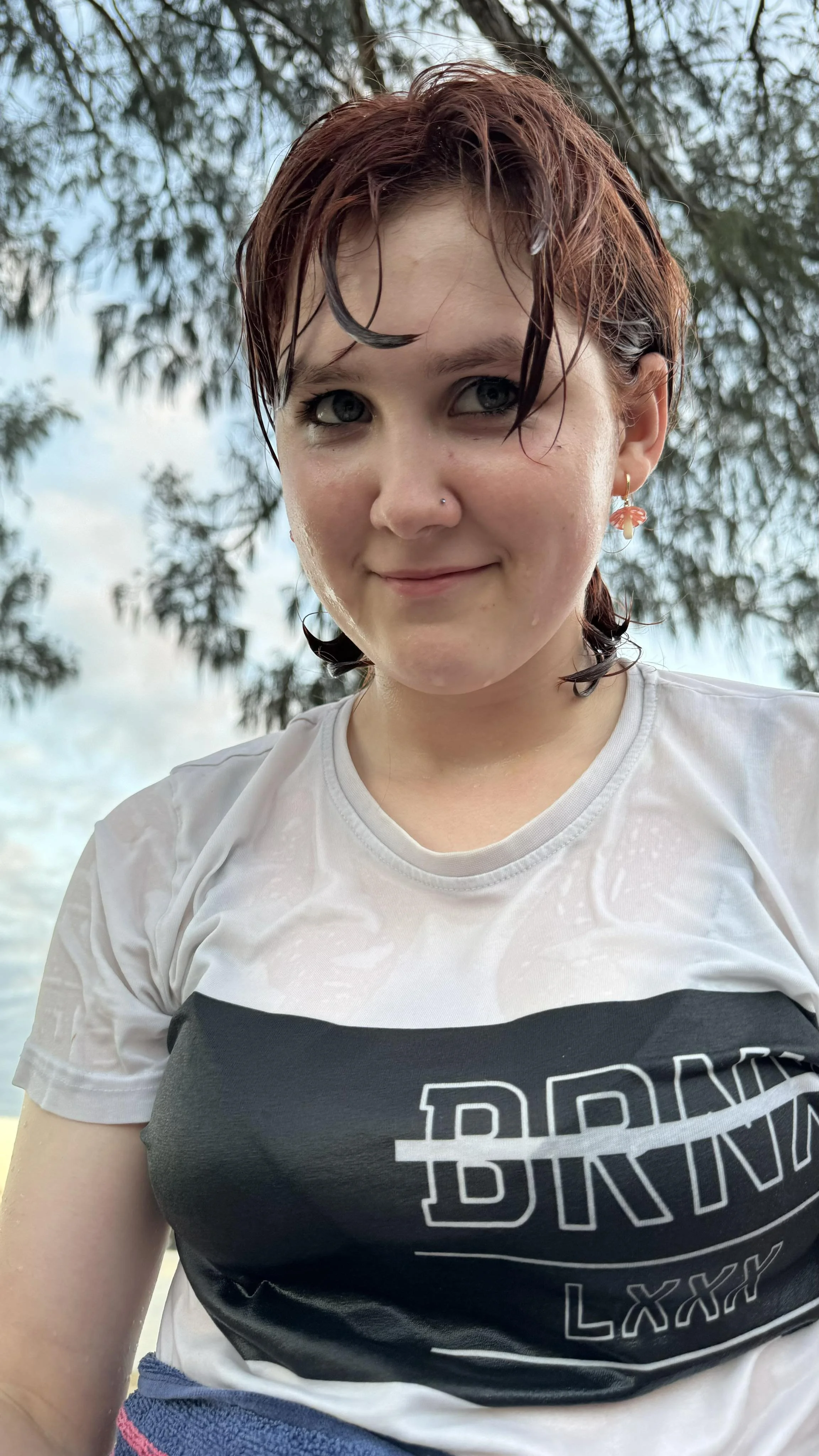 A young woman with short, wet, reddish-brown hair outdoors, wearing a white shirt with black text, earring, and a nose piercing.