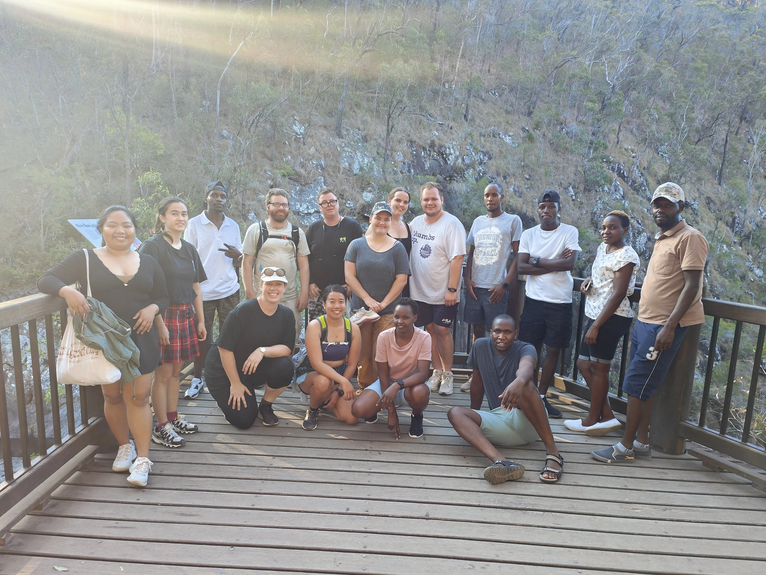 Group of people standing and sitting on a wooden viewing platform outdoors with a background of trees and rocky terrain, sunlight shining from the top left.