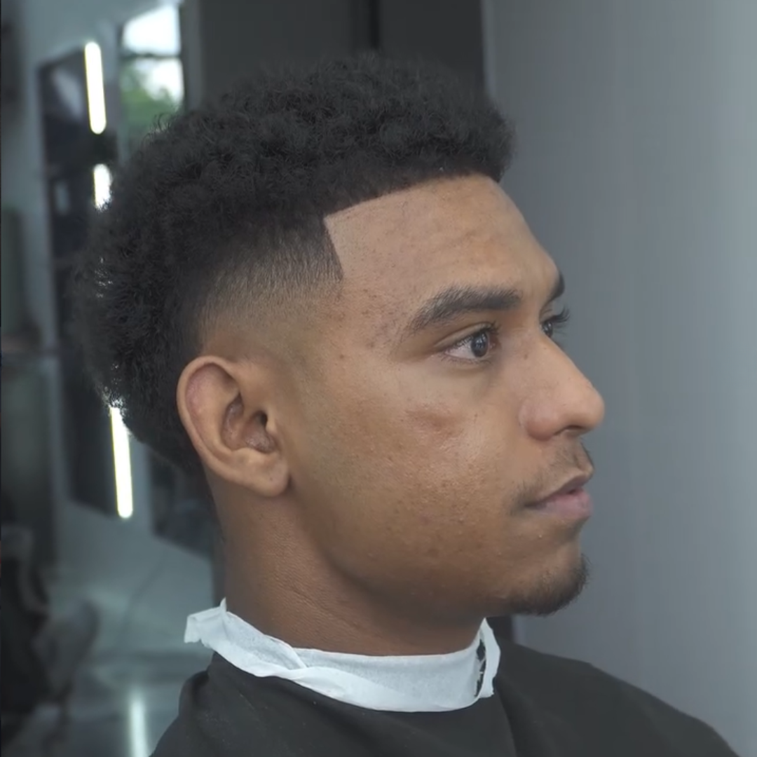 A young man in a barber's chair after receiving a fresh haircut with a fade hairstyle, wearing a smock and cape.