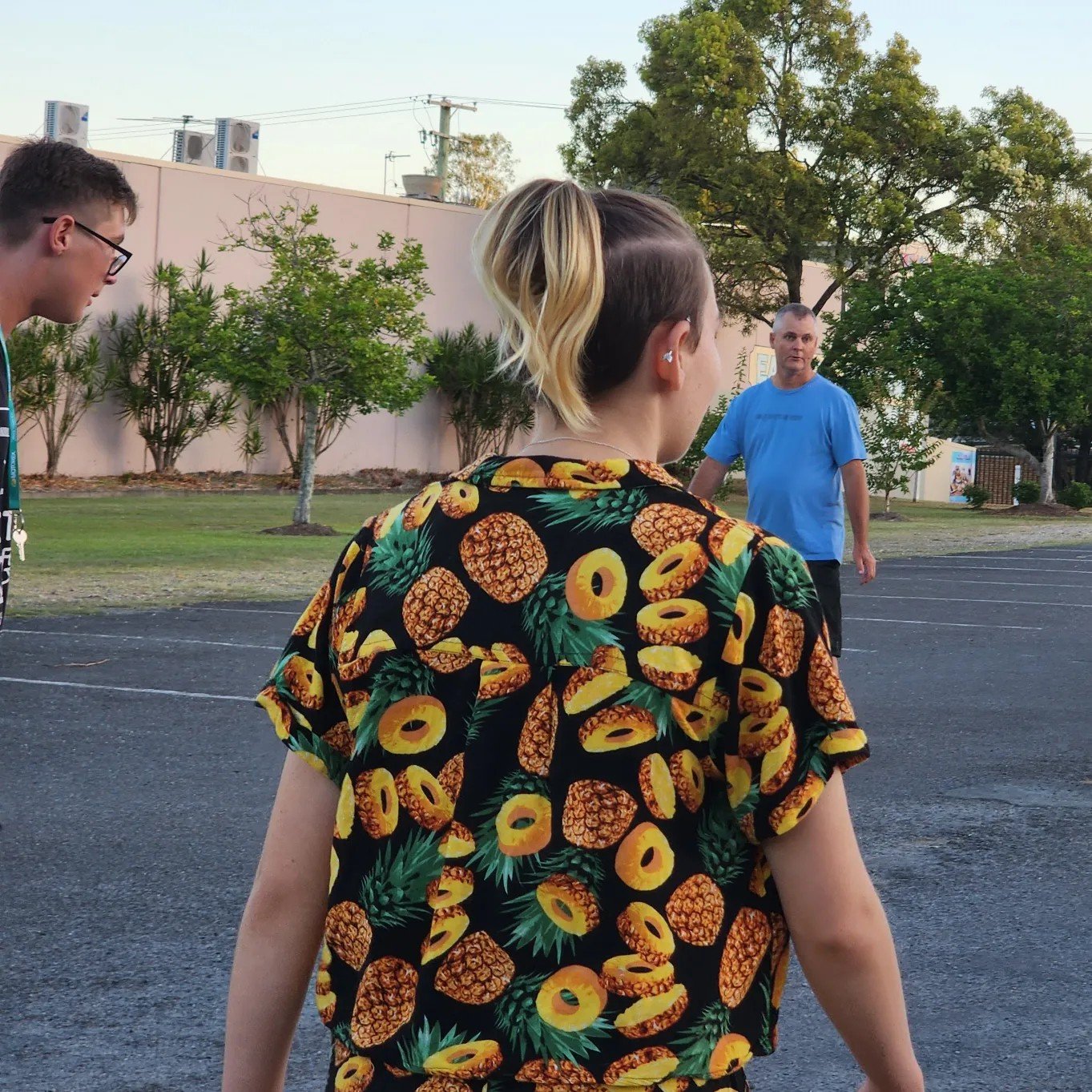 People standing outdoors in a parking lot with trees and a building in the background. One person wears a black shirt with a pineapple and pineapple slice pattern.