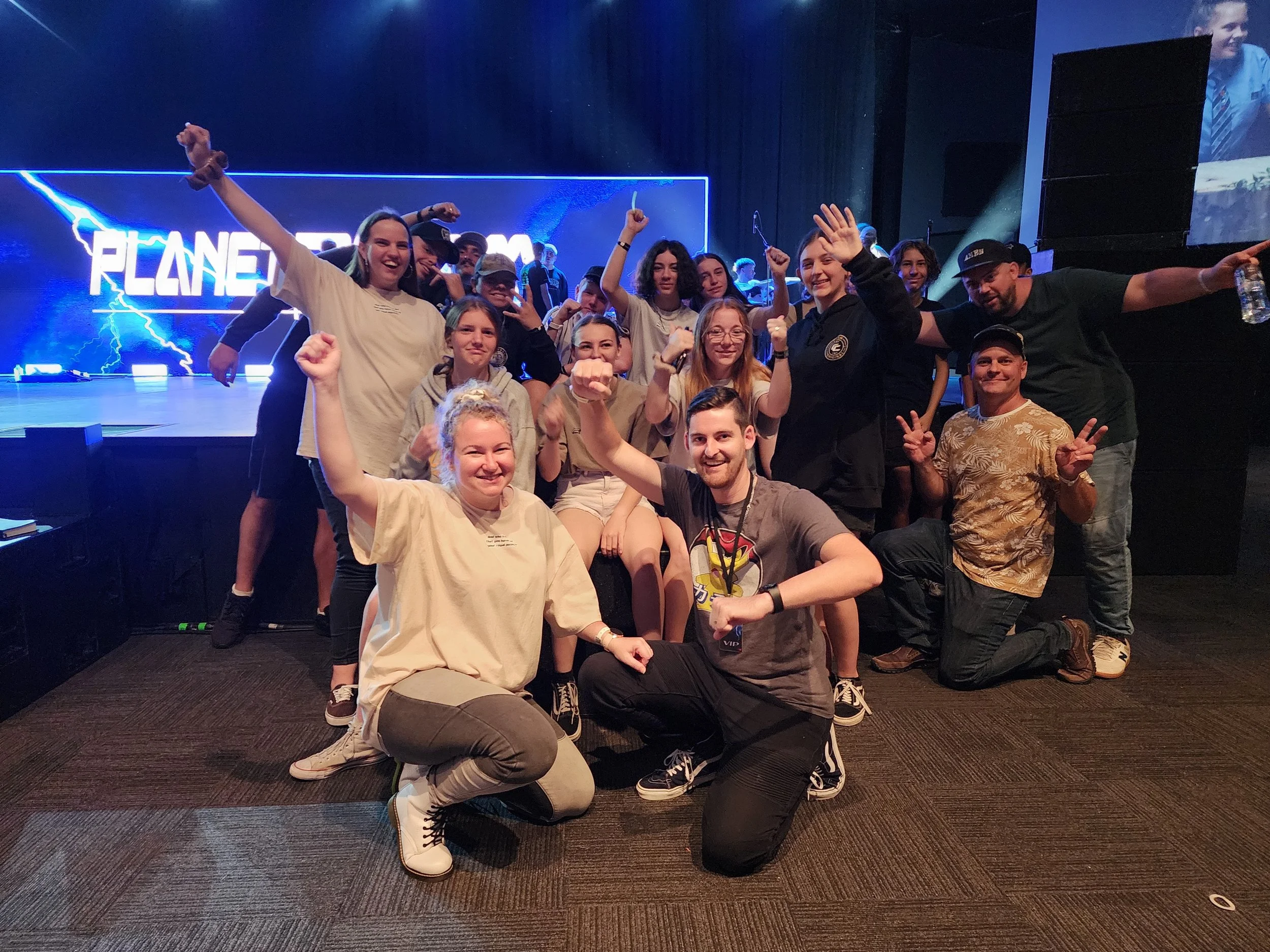 Group of young people celebrating on stage with a large screen behind them displaying the word 'PLANET SHAKERS' surrounded by lightning graphics.