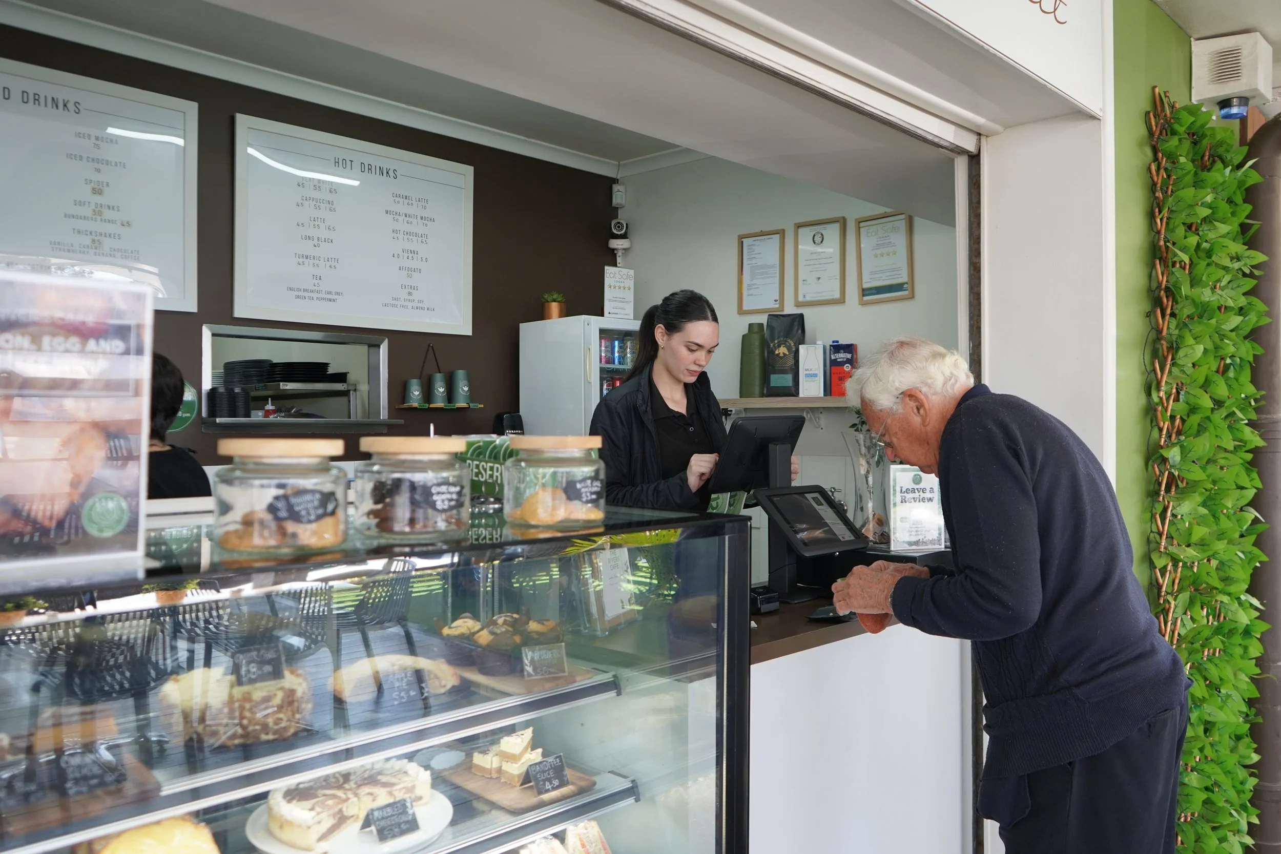 A man paying at a cafe counter while a barista takes his order. The display case shows various cakes and pastries. The menu boards are visible in the background.