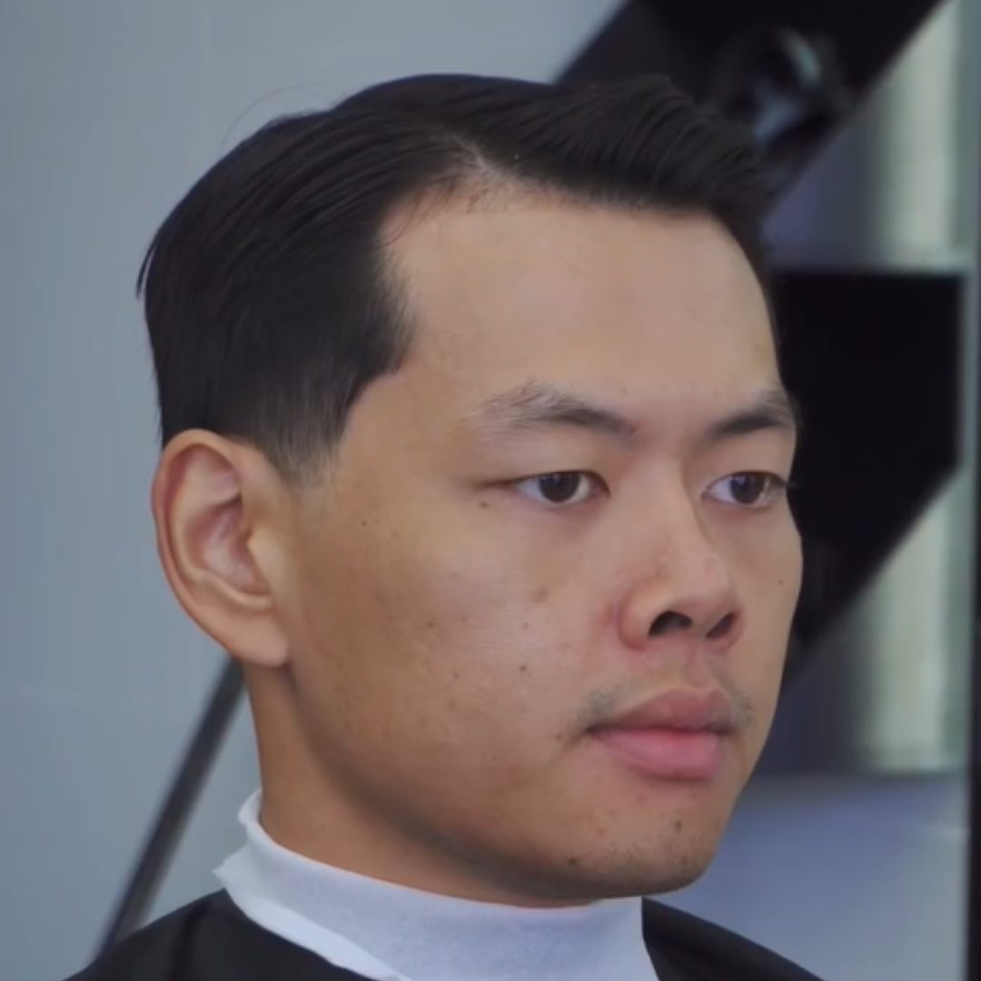 Close-up of a man with short black hair, sitting in a barber's chair, getting a haircut.