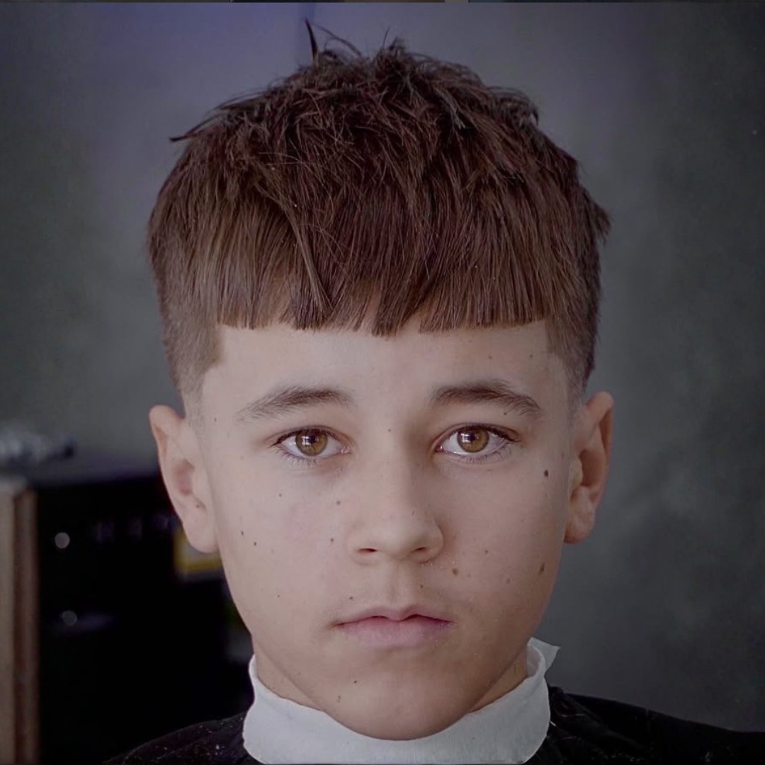 Close-up of a young boy with brown hair and brown eyes, sitting in what appears to be a barber's or salon chair.