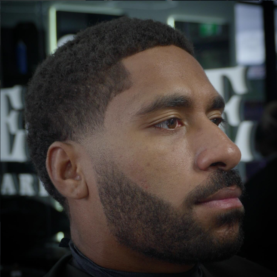 Close-up of a man with short, curly hair and a beard, sitting in a barber shop or salon.