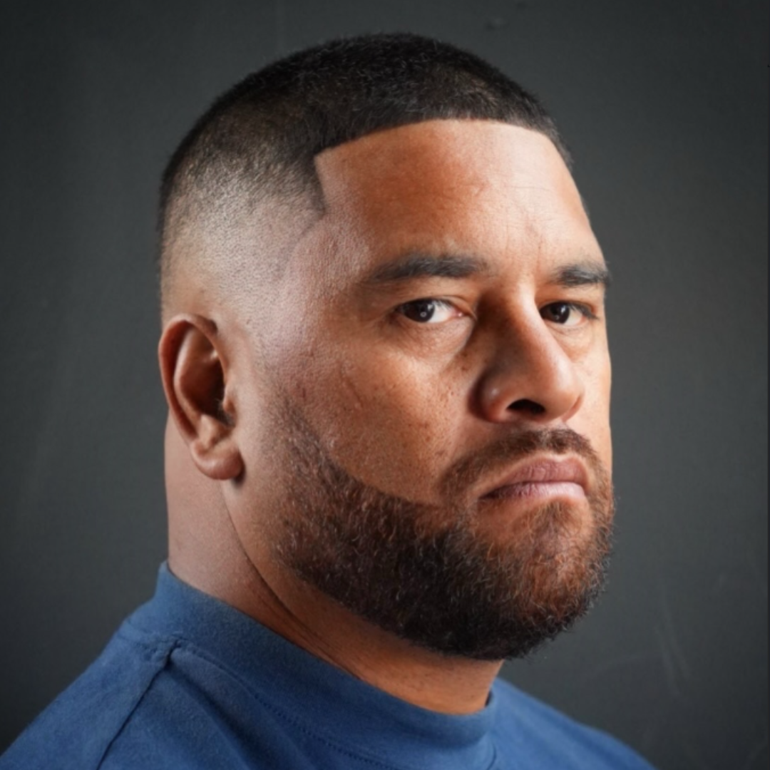 Close-up portrait of a man with a serious expression, short hair, beard, and wearing a blue shirt, against a dark background.