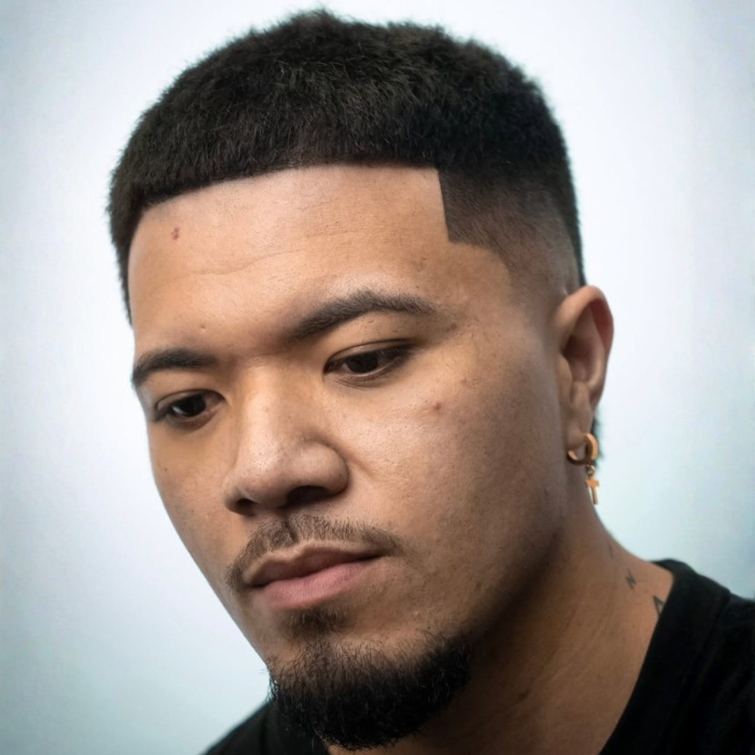 Close-up portrait of a young man with a short hairstyle, earring, and facial hair, looking down.