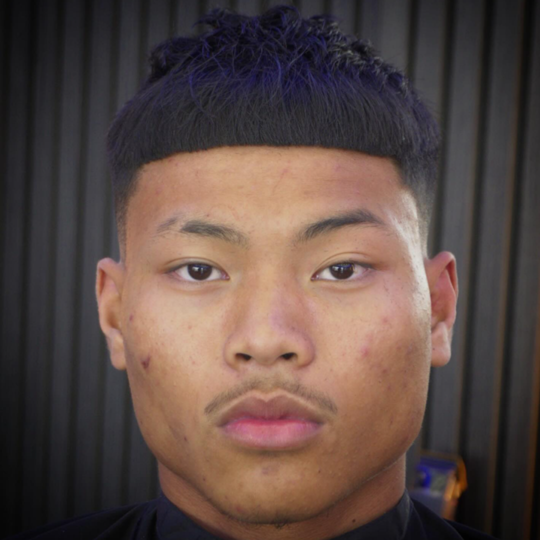 Close-up of a young man with a short, dark hairstyle, acne on his face, and neutral expression, standing in front of a dark, vertically paneled wall.