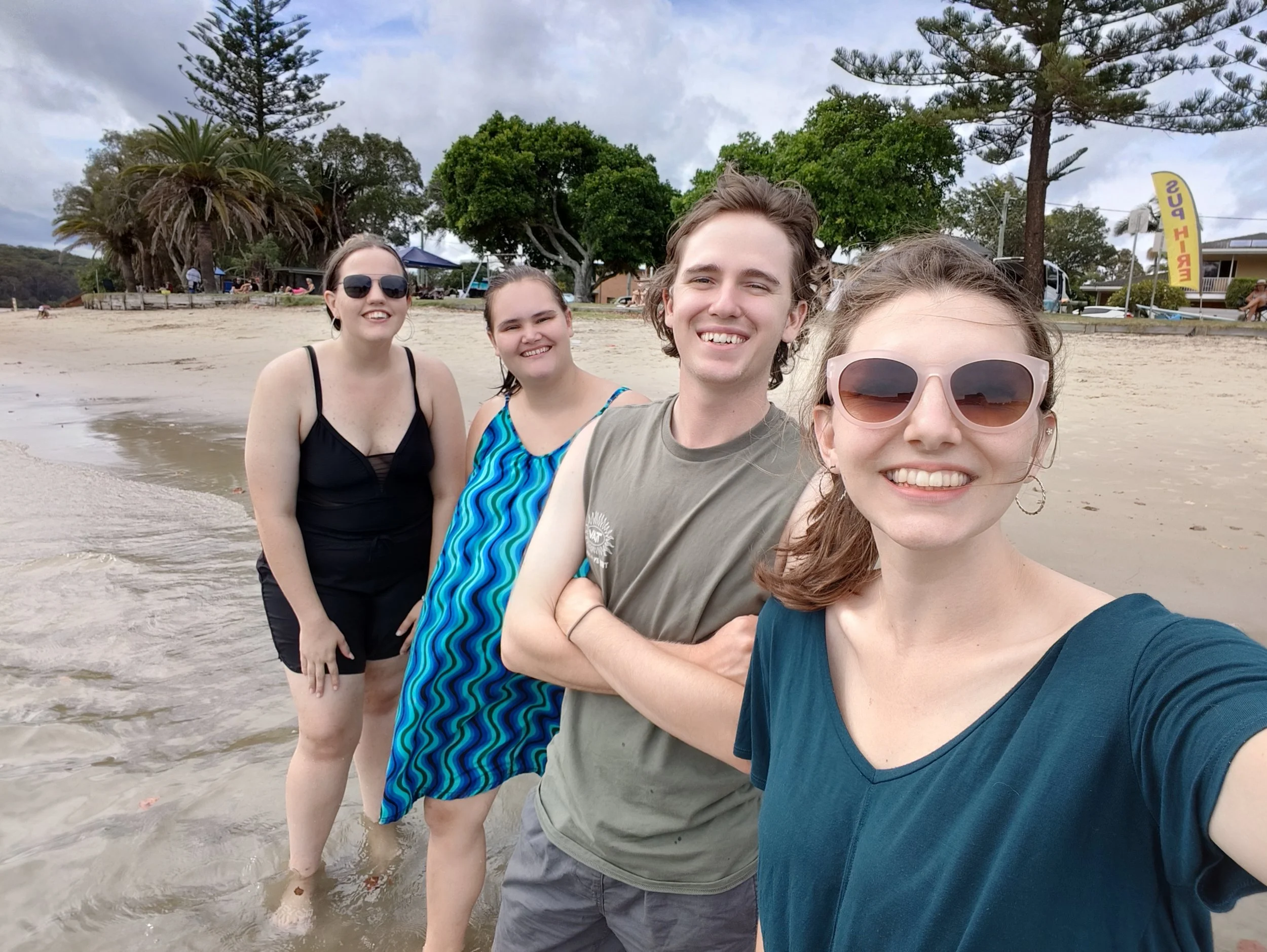 Four friends smiling and taking a selfie on a beach with trees and umbrellas in the background.