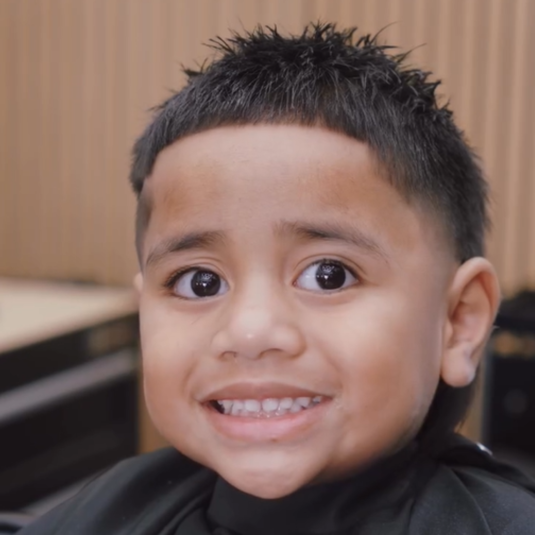 A young boy with a short, spiky haircut smiling in what appears to be a barber shop.