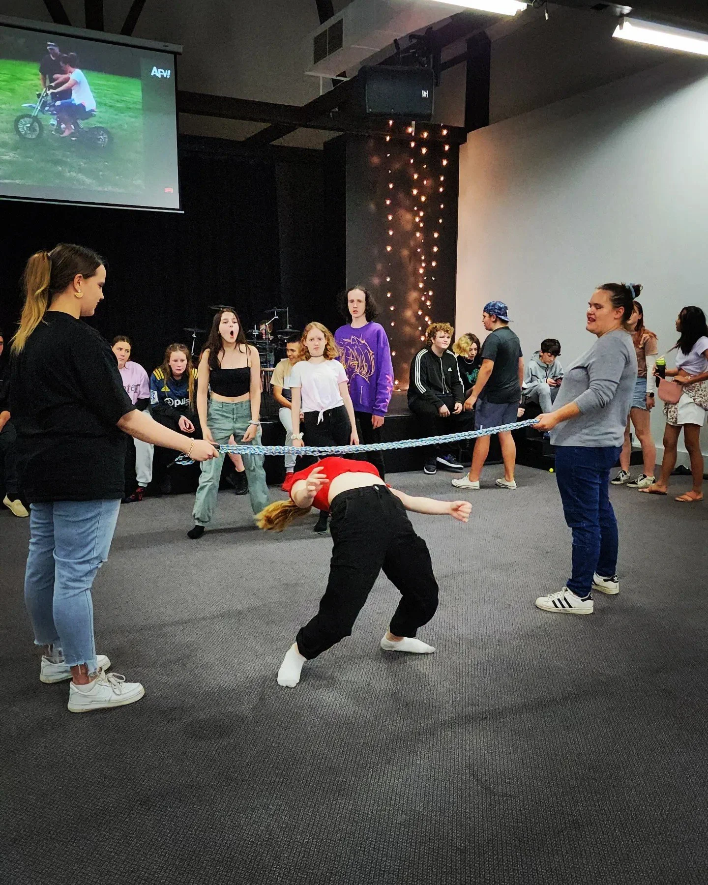 A group of young people playing a game of limbo in a large indoor space, with some onlookers in the background.