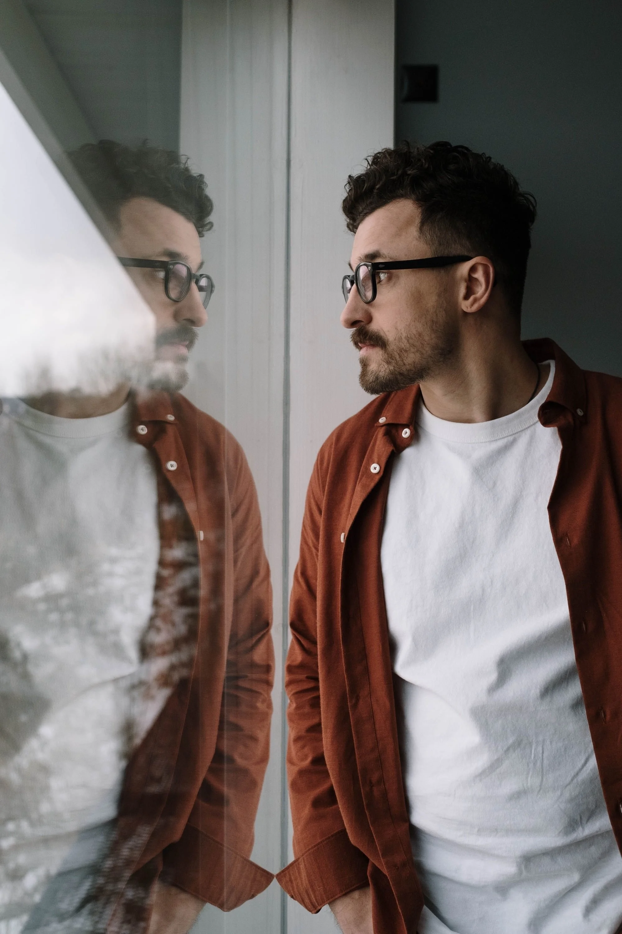 Man in a rust colored shirt looking thoughtfully out a window, his reflection visible in the glass.