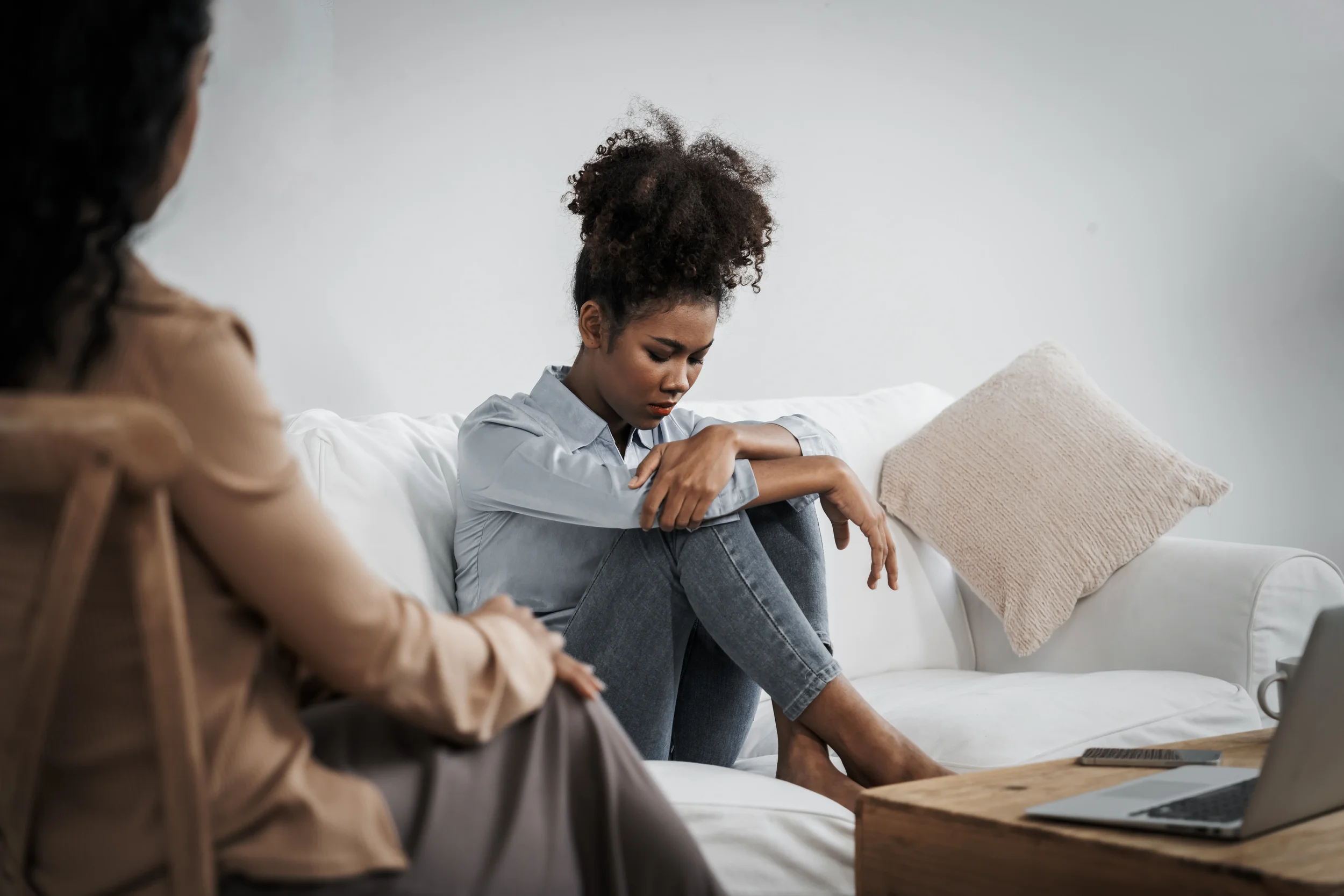 Young woman sitting on white couch with knees drawn up during therapy session, therapist visible in foreground