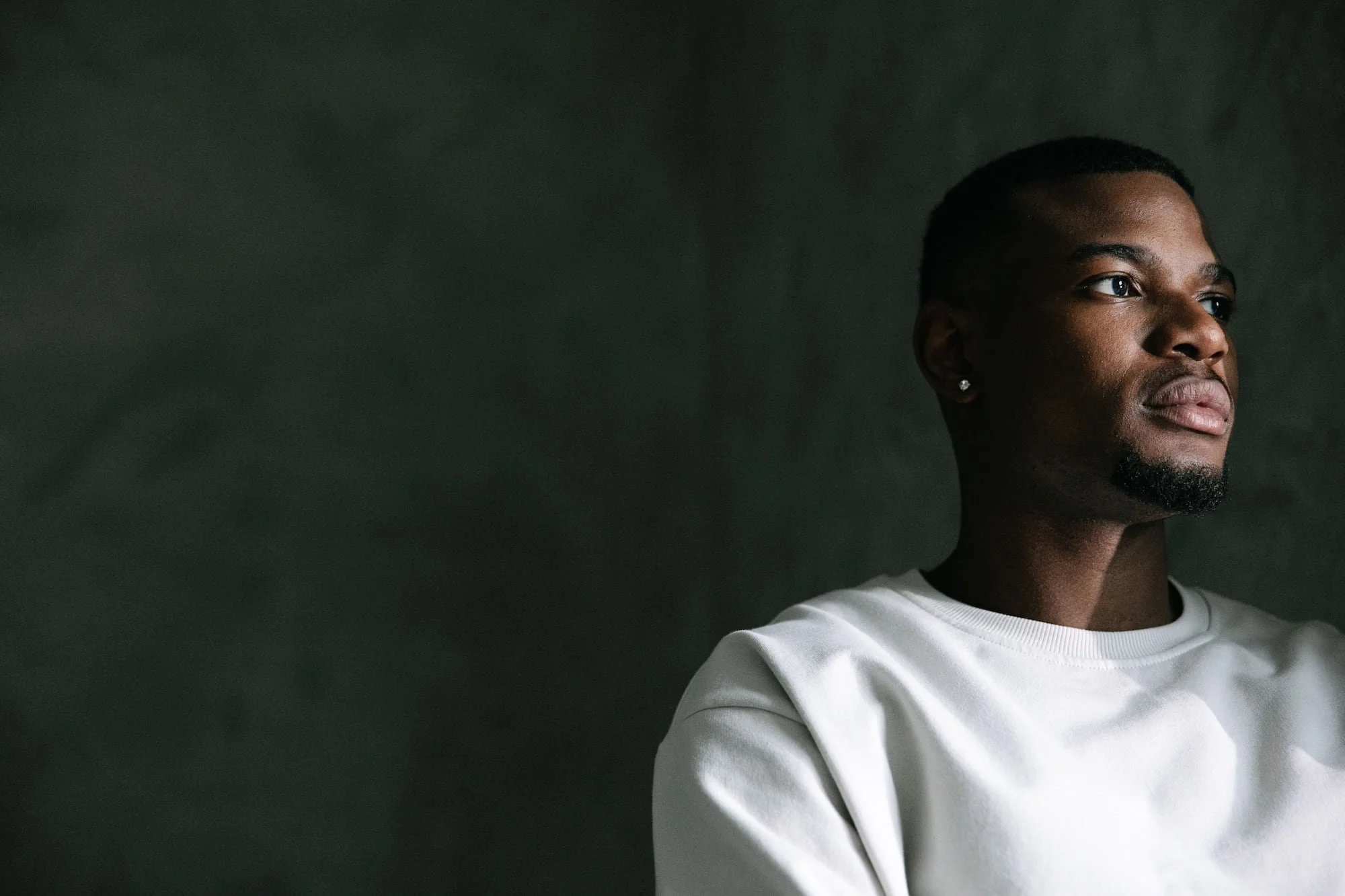 A man with a short beard and stud earring, wearing a white crewneck sweatshirt, gazes thoughtfully to the side while dramatically lit against a dark green background.