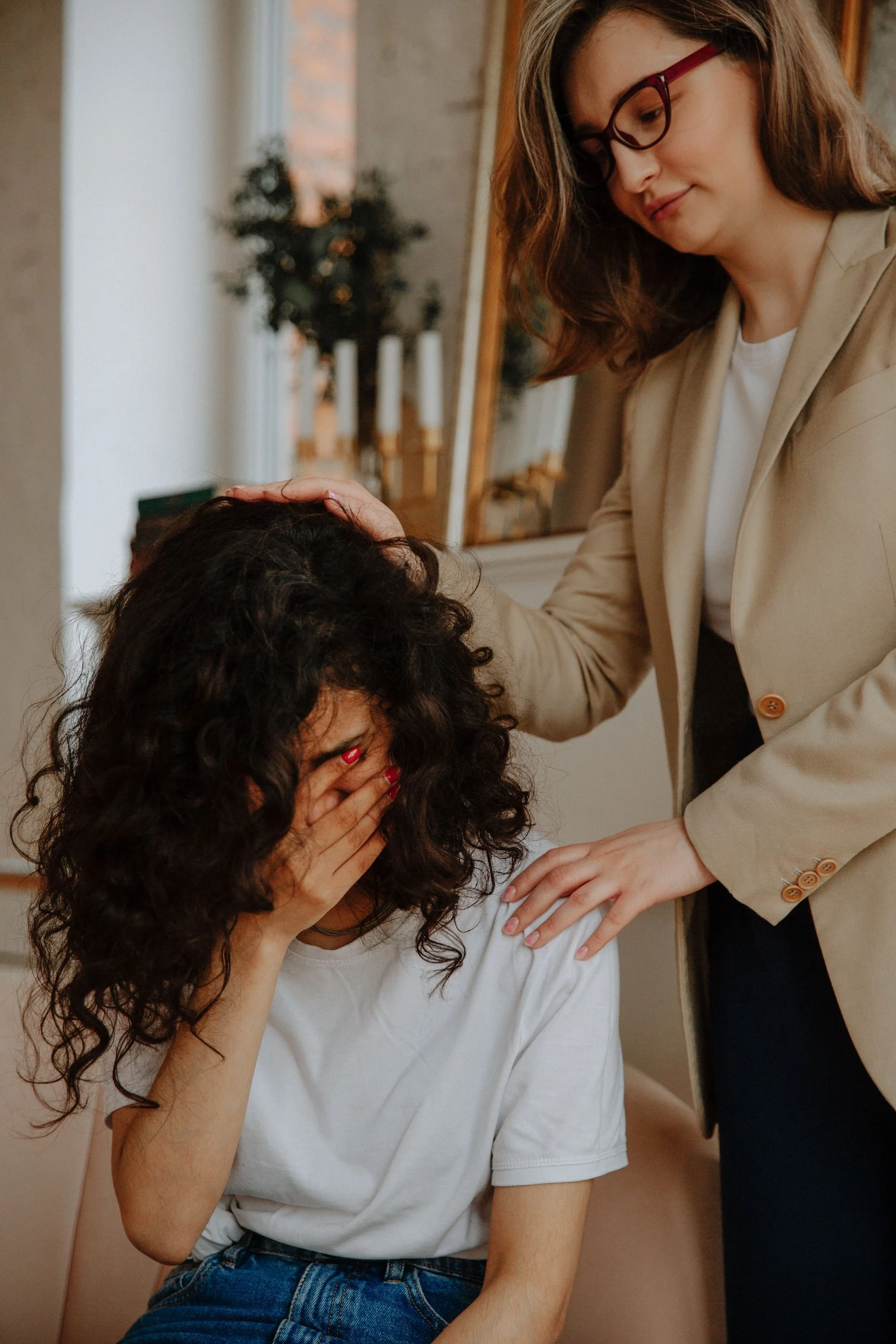 A therapist in a beige blazer gently comforts a young woman with curly hair who is covering her face with her hand, resting a supportive hand on her shoulder and head.
