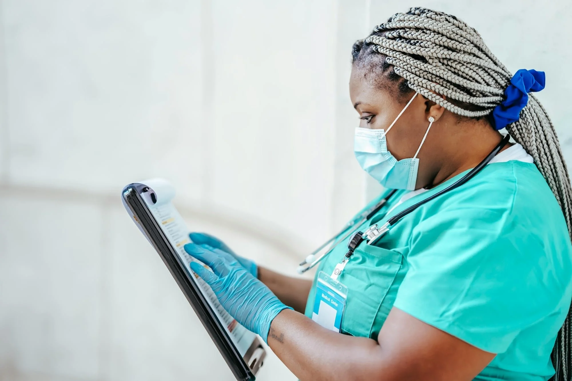 A Black female nurse in teal scrubs, gloves, and a face mask reviews a patient chart on a clipboard, representing the diverse healthcare workers and professionals who seek support through multicultural counseling in North Riverside, Illinois.