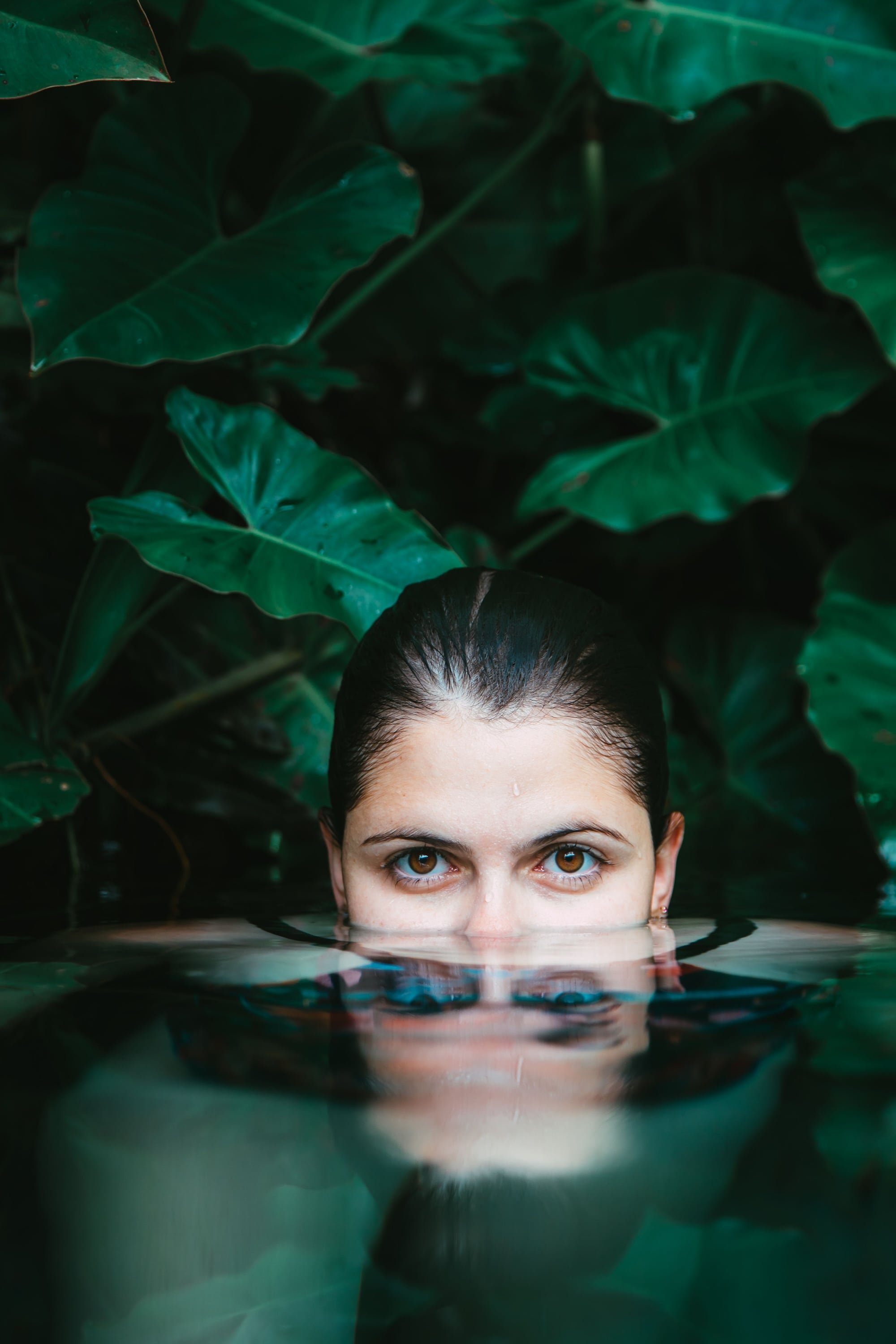 Woman submerged to eye-level in dark water, surrounded by large green tropical leaves, calm and focused gaze