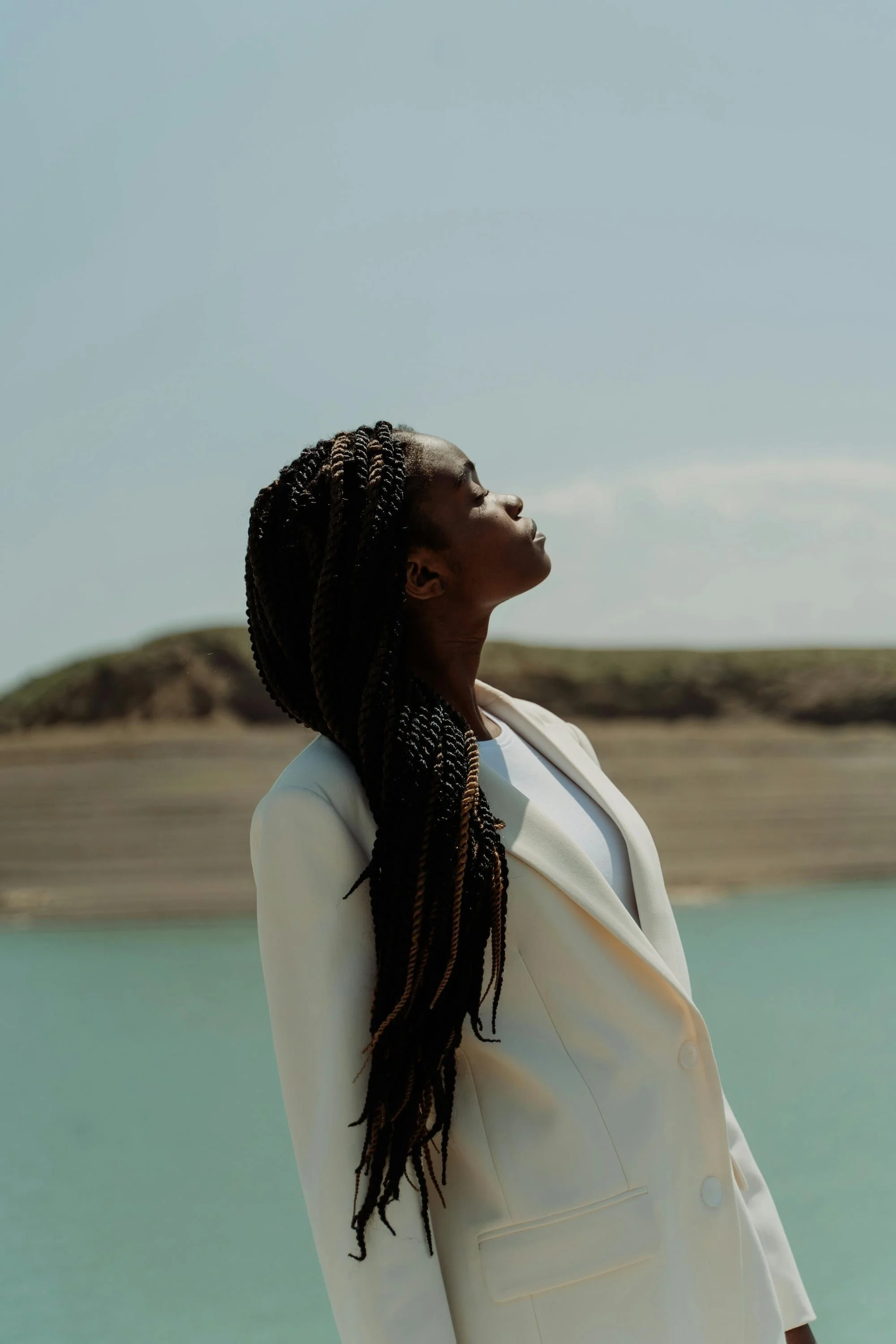 A young woman with long braided twists tilts her head back toward the open sky, wearing a white double-breasted blazer, standing beside a turquoise body of water with sandy hills in the background.