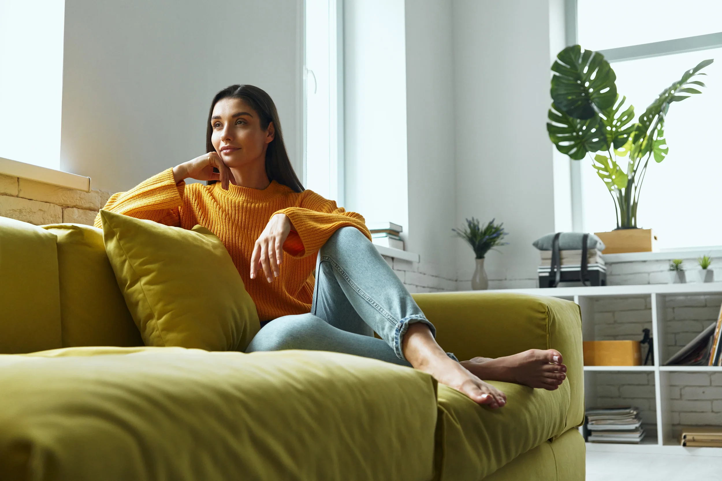 Woman in orange sweater sitting thoughtfully on green couch in bright, modern living room with plants