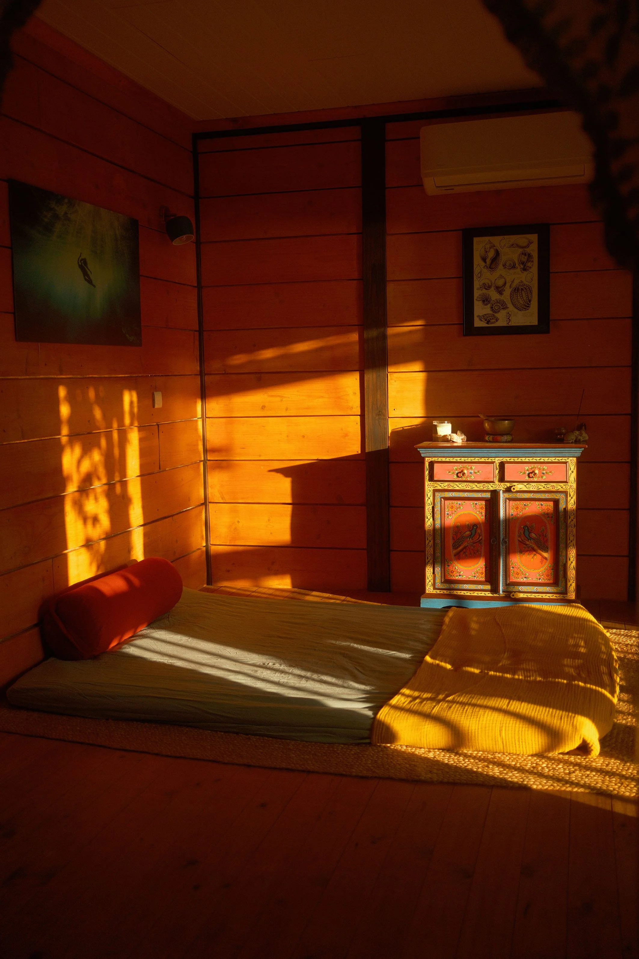 Une chambre avec des murs en bois, un lit au sol avec un oreiller cylindrique rouge, une couverture jaune, une petite table colorée avec des objets dessus, et deux portraits encadrés accrochés au mur. La pièce est éclairée par des rayons de soleil.