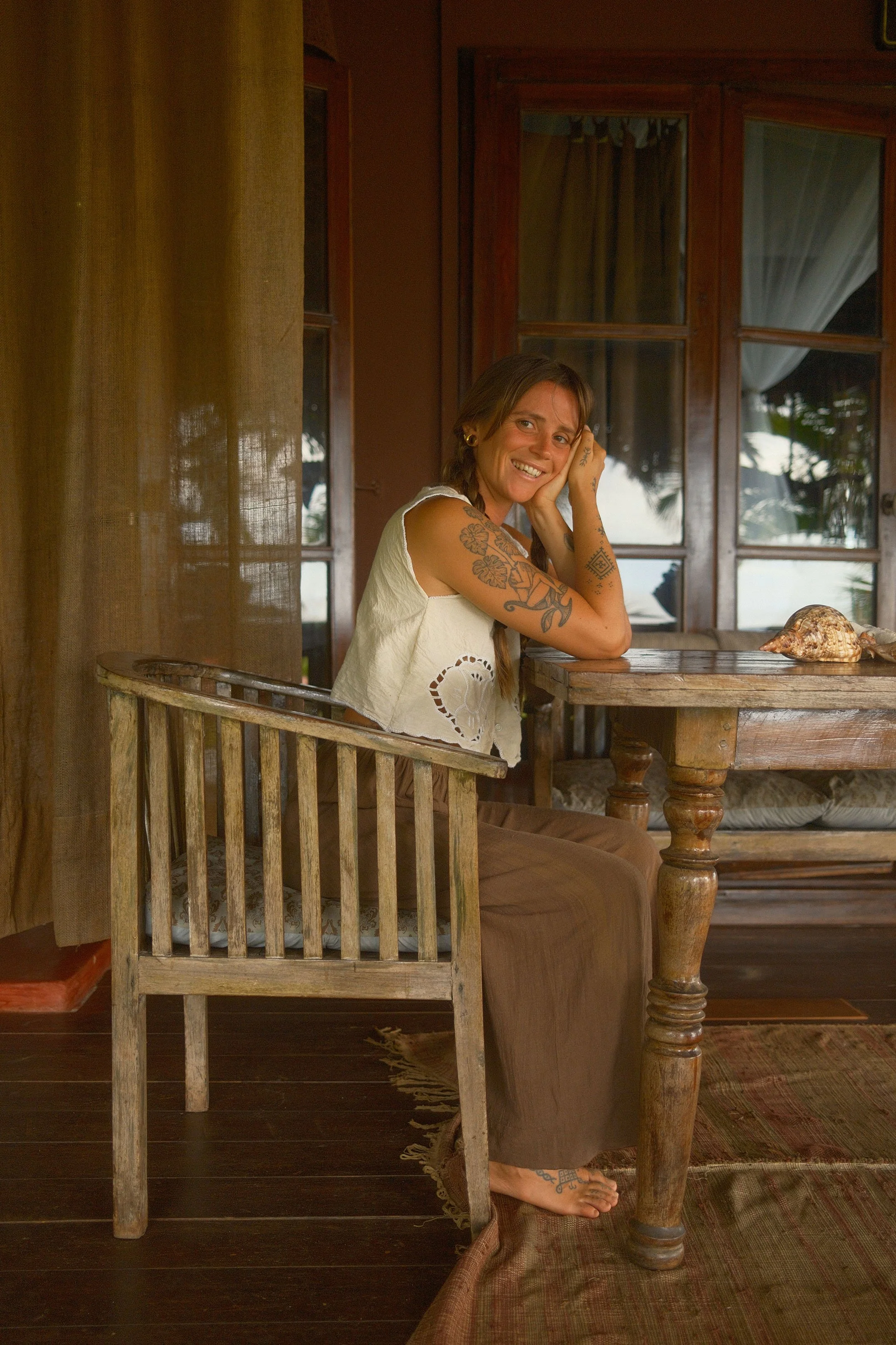 Une femme avec des tatouages, assise à une vieille table en bois, dans une pièce en bois, souriant et posant sa tête sur sa main. Une coquille de coquillage est sur la table devant elle.