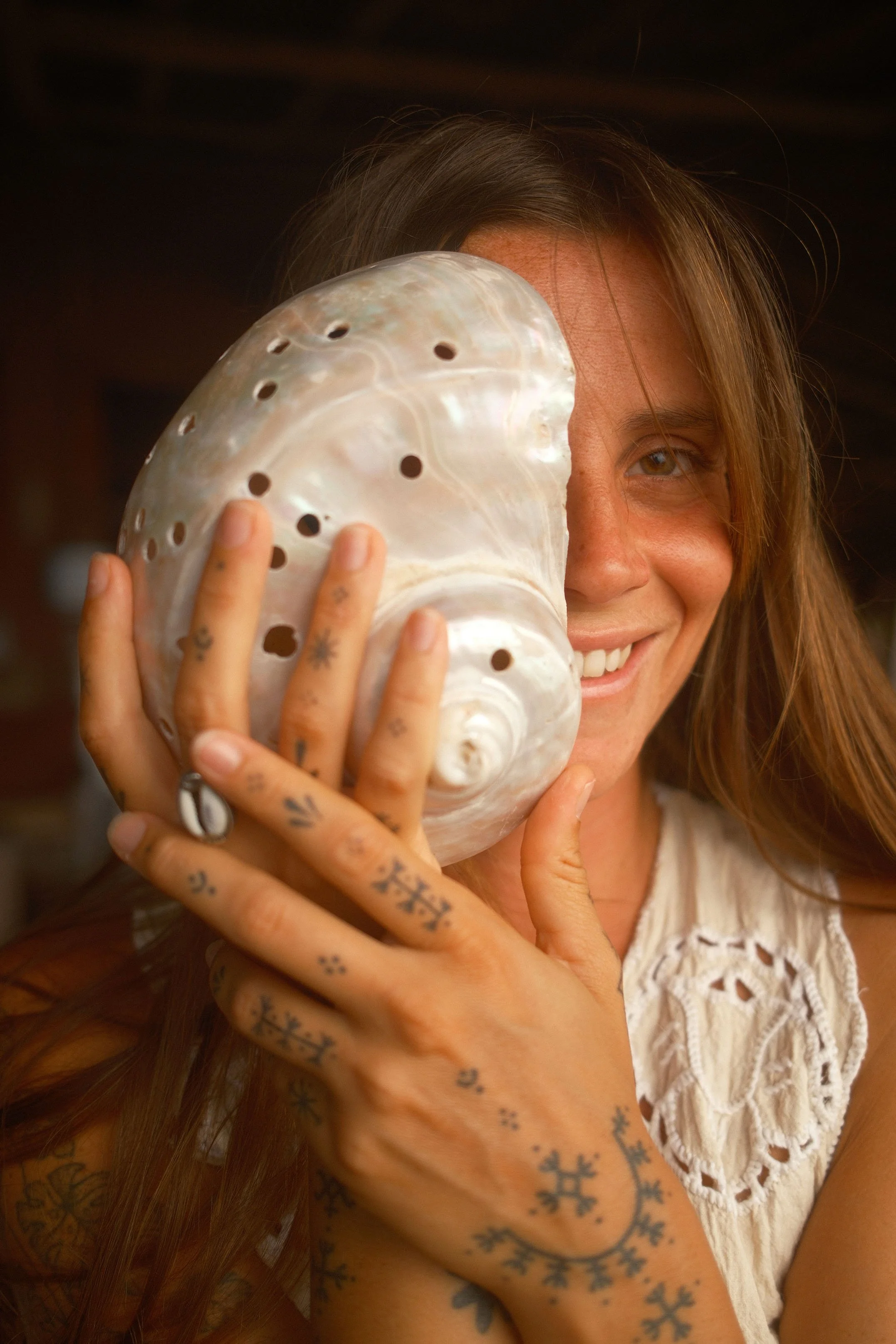 Une femme souriante tenant un coquillage blanc avec des trous près de son œil, avec des tatouages visibles sur ses mains et bras, portant une blouse blanche en dentelle.